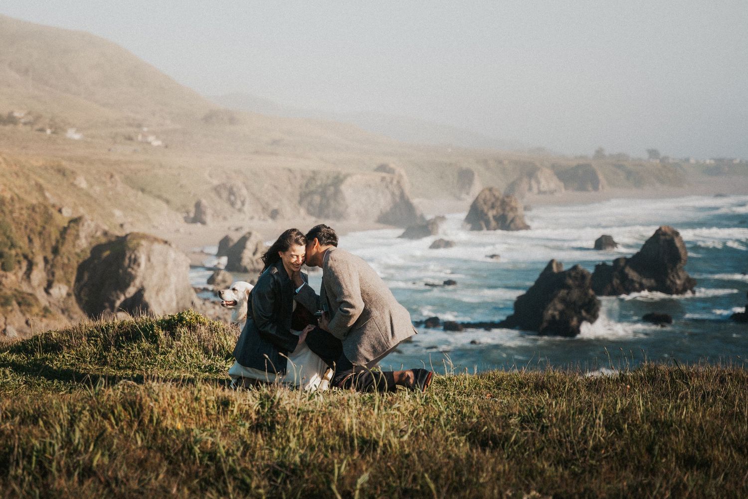 Couple sitting together on coastal cliffside overlooking rocky Pacific Ocean shoreline with misty hills in background.