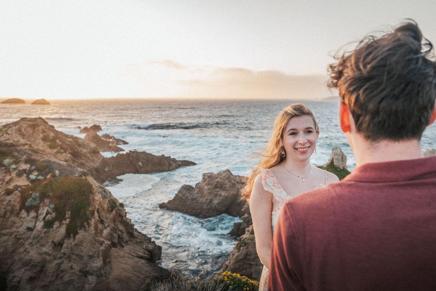 A smiling blonde woman poses on rocky coastal cliffs at sunset with ocean waves in the background.