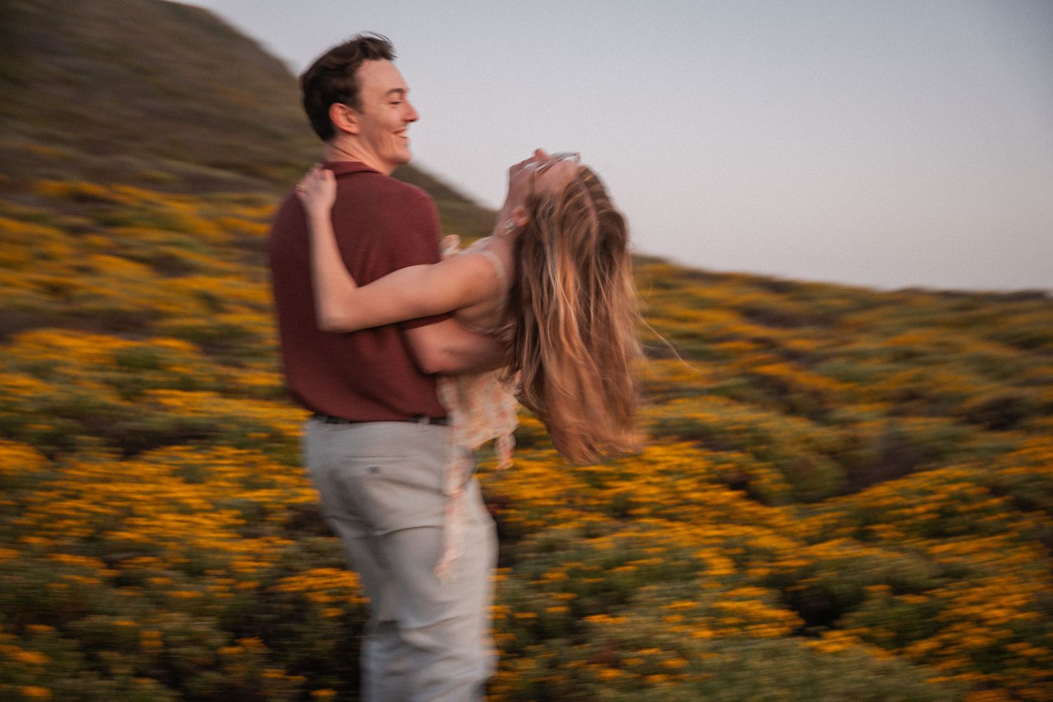 Couple joyfully embracing in a vibrant wildflower field, man lifting woman while both laugh outdoors.