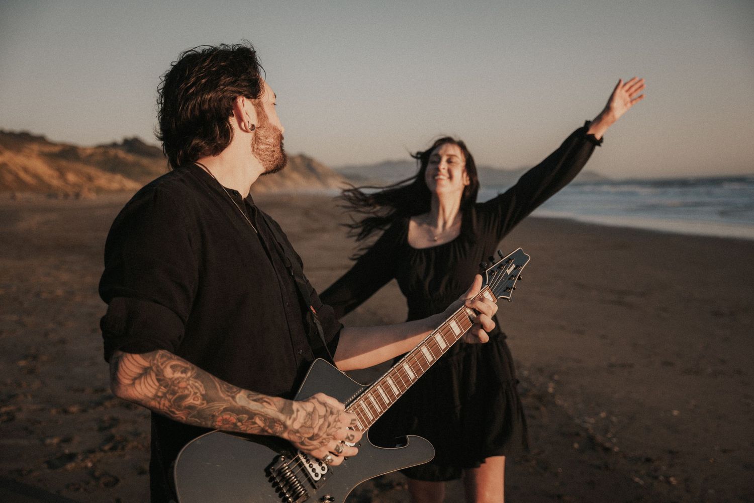 Man with tattoos playing electric guitar on beach at sunset while woman in black dress dances with arms raised.