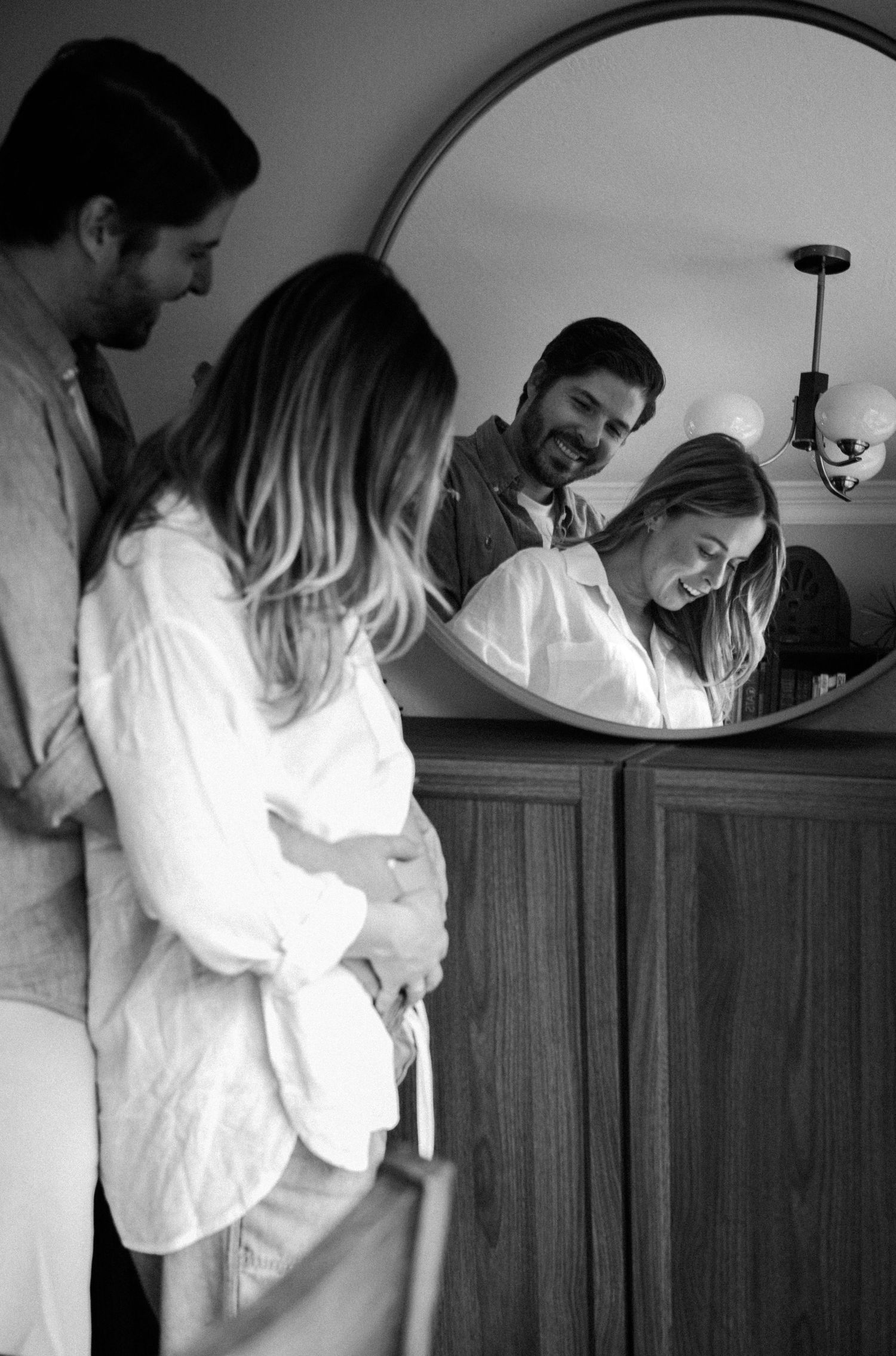 Black and white photo of a couple laughing together, reflected in a round mirror above a wooden dresser.