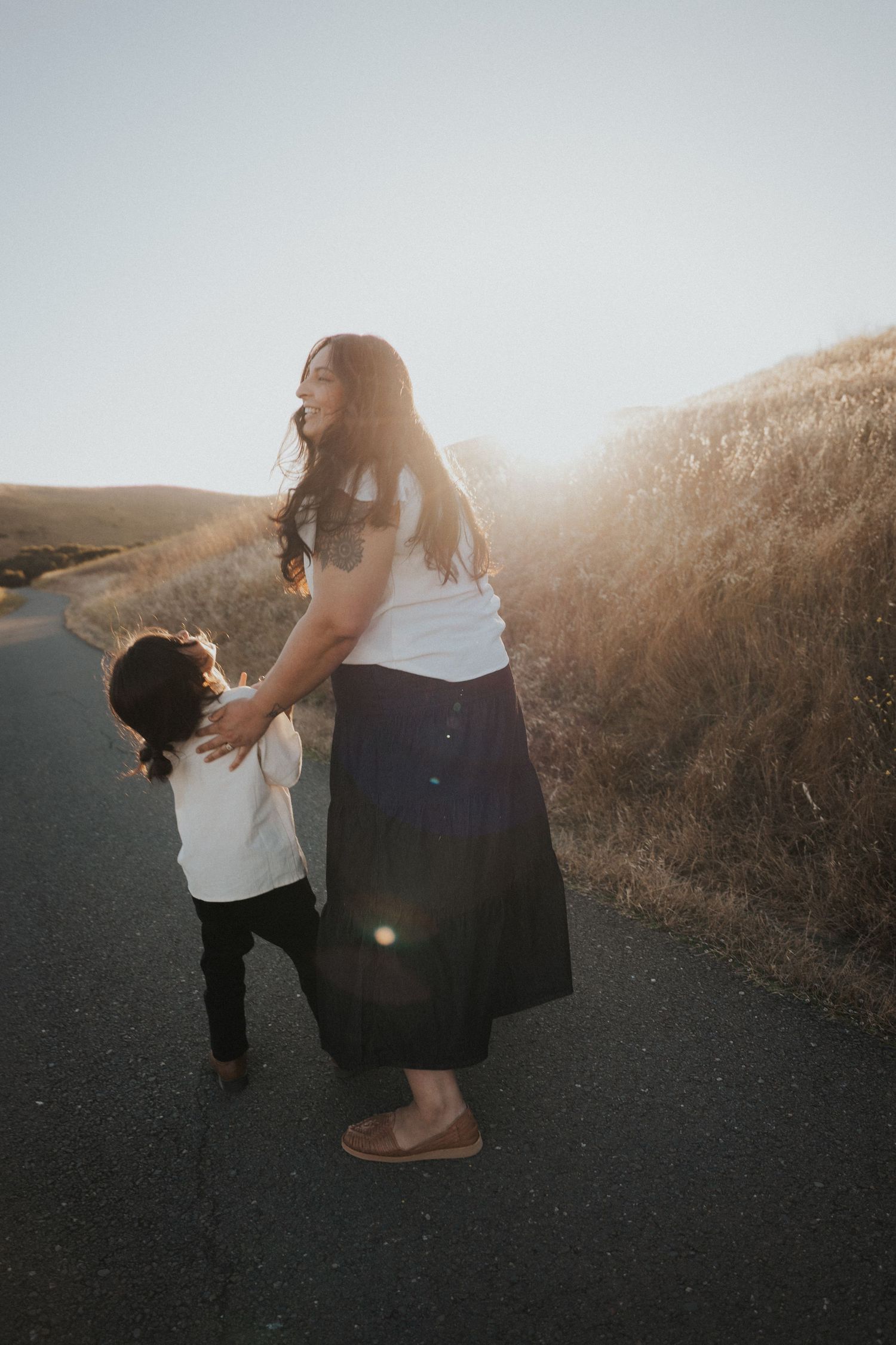 Mother and child holding hands on a sunlit rural road surrounded by golden hills at sunset.