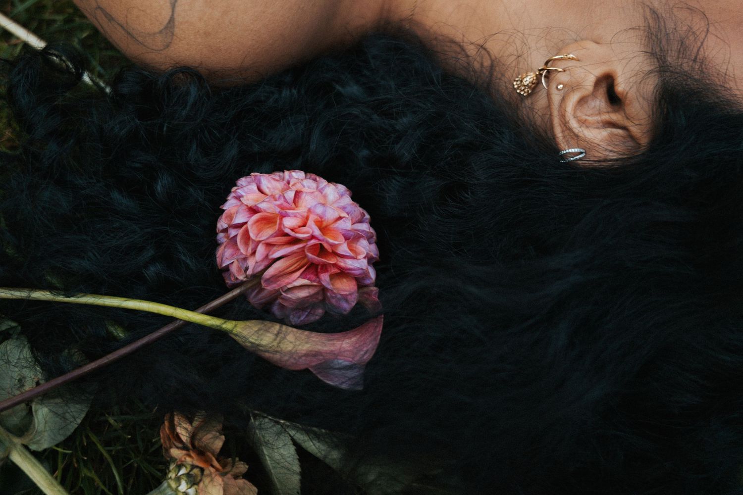Person with dark hair lying down holding a pink dahlia flower surrounded by lush green foliage.