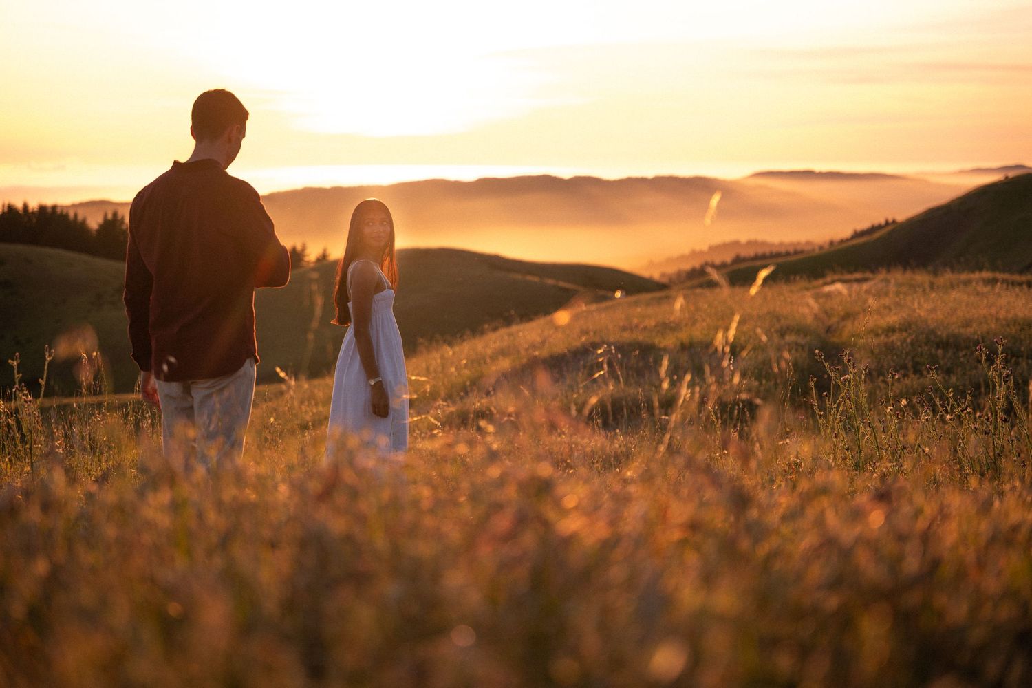 Couple standing in golden field at sunset with misty hills and warm glowing sky in the background.