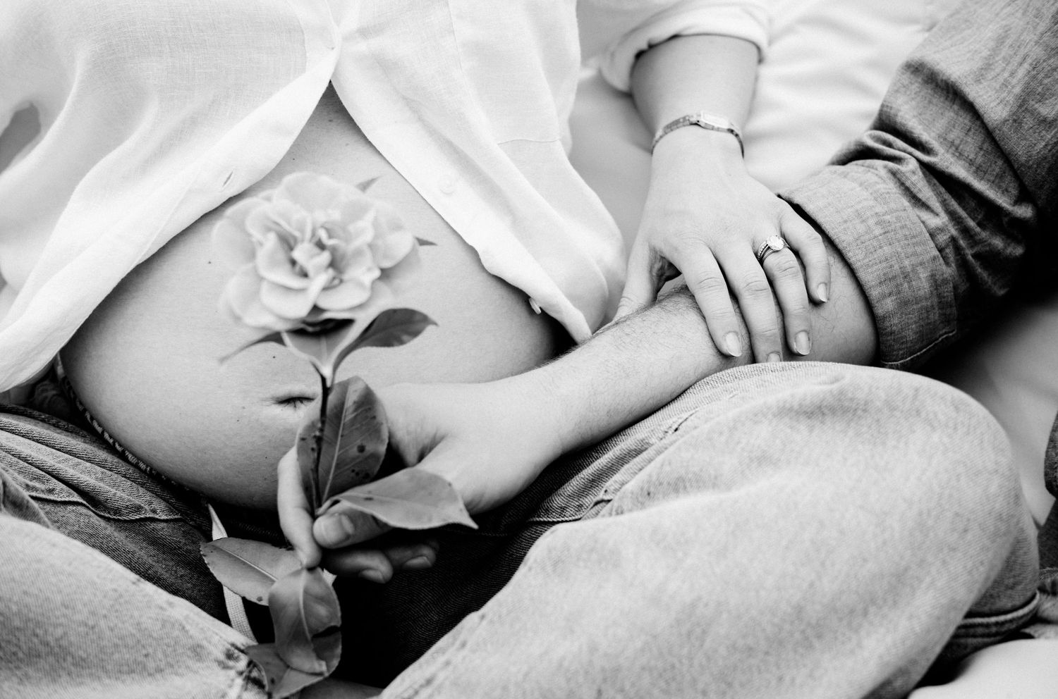 Black and white photo of pregnant woman's bare belly with a rose flower, partner's hand resting on her side.