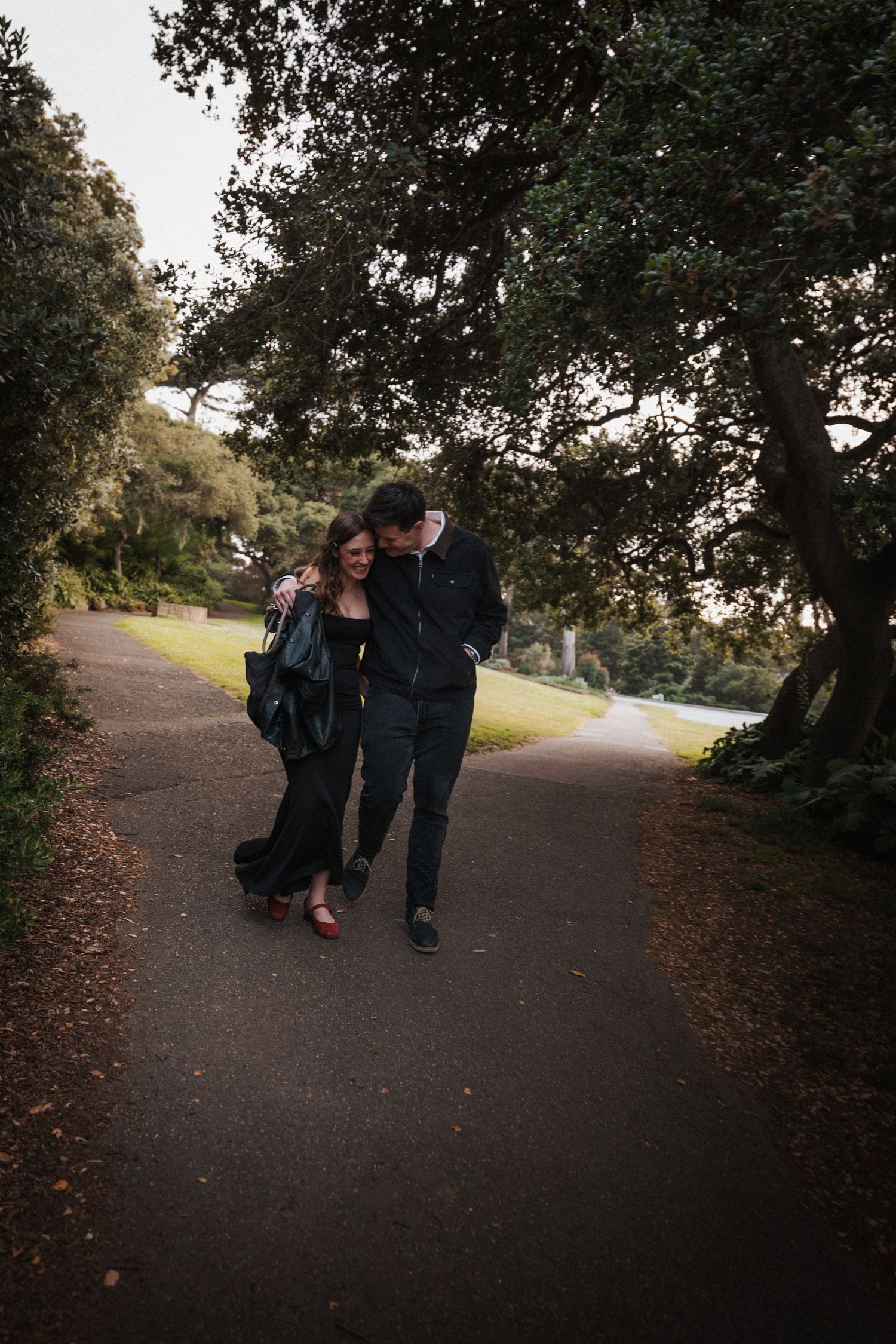 A couple walks arm in arm down a tree-lined park path, both dressed in black, sharing a joyful moment.