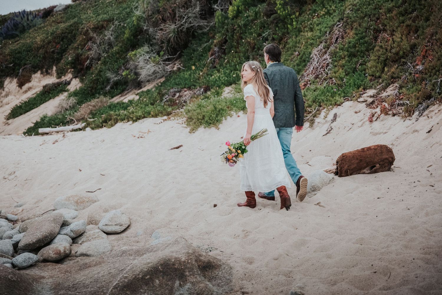 Bride and groom walking on sandy beach holding bouquet with dog nearby, green cliffs in background.