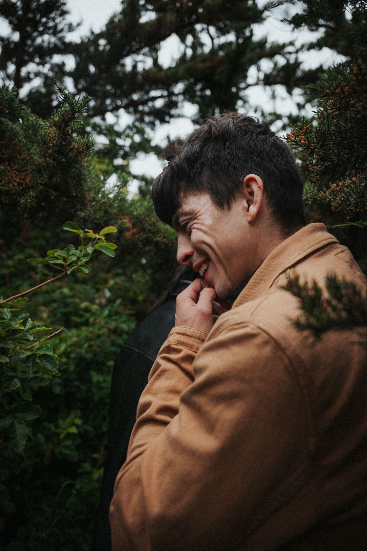 Man in tan jacket laughing candidly among lush green cypress trees outdoors.
