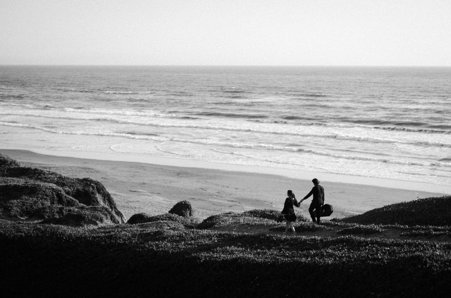 Silhouette of couple holding hands walking toward ocean waves on rocky beach in black and white.