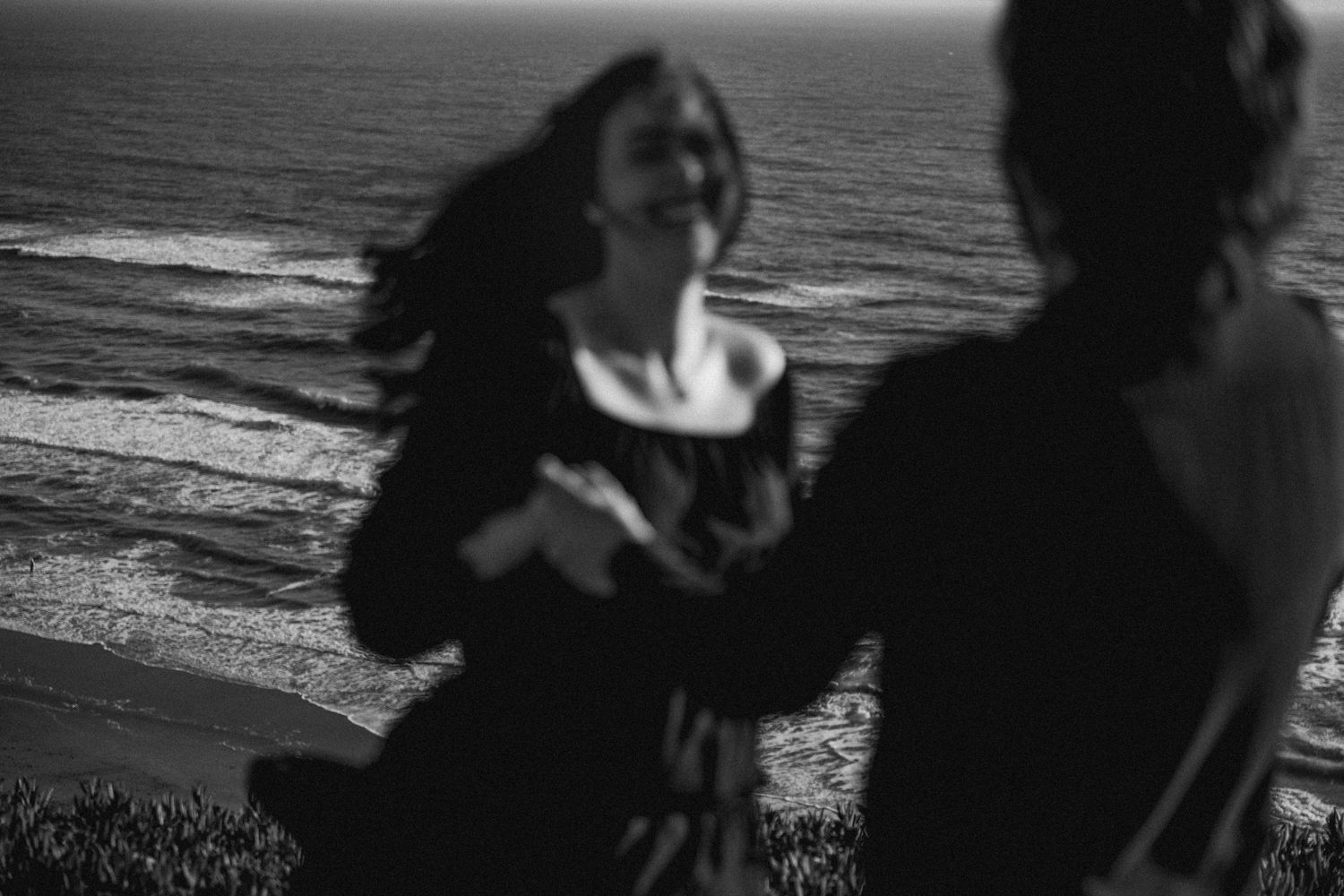 Two women laugh and dance together on a coastal cliff overlooking ocean waves in black and white.