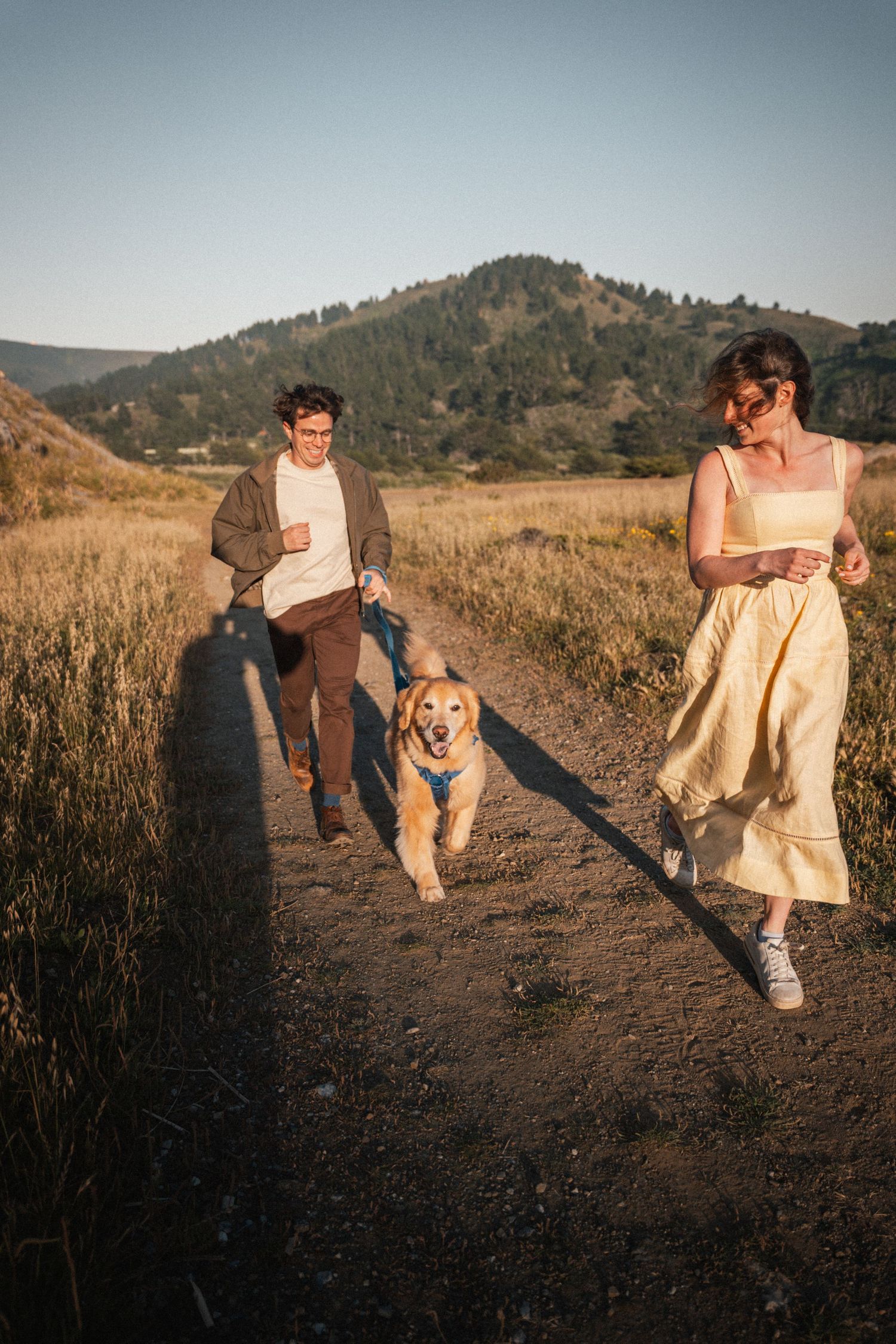 Couple running with golden retriever on dirt path through golden fields with mountain backdrop at sunset.