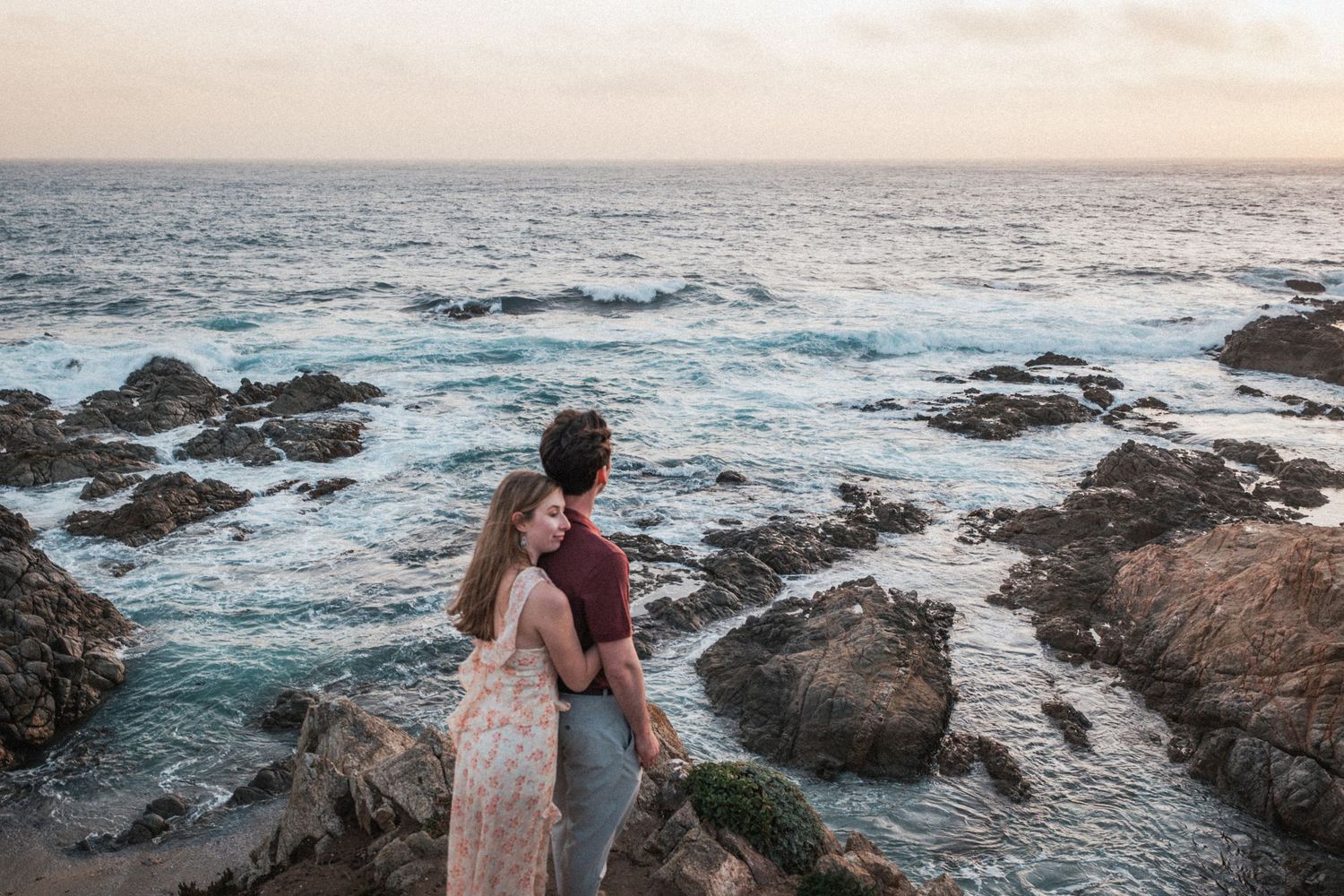 Couple embracing on rocky coastal cliffs at sunset overlooking the ocean.