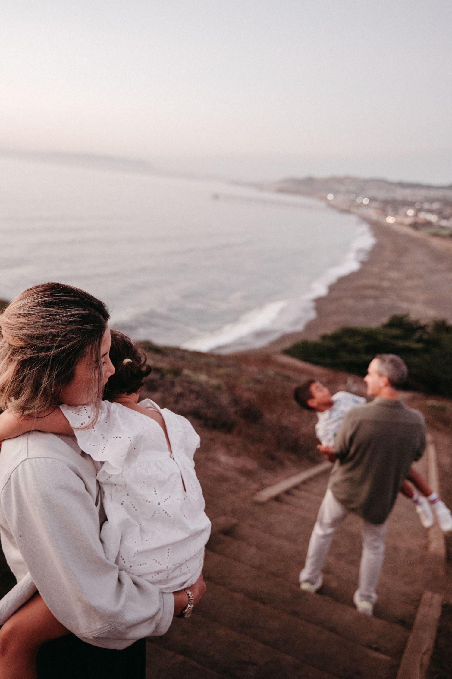 Woman holding child on coastal overlook at dusk with ocean view.