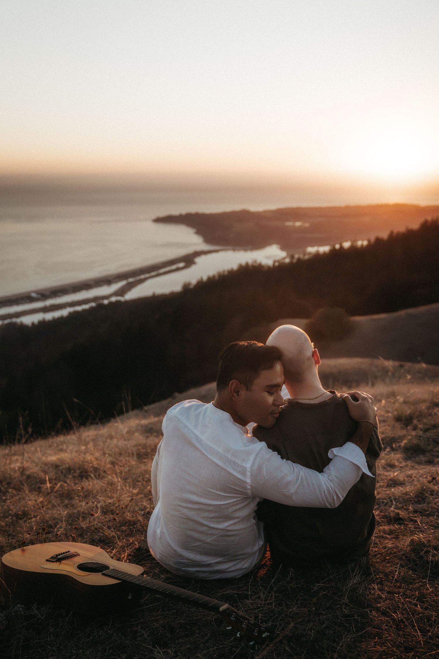 Two people embrace on a hillside at sunset with a guitar nearby, overlooking a scenic coastal landscape.