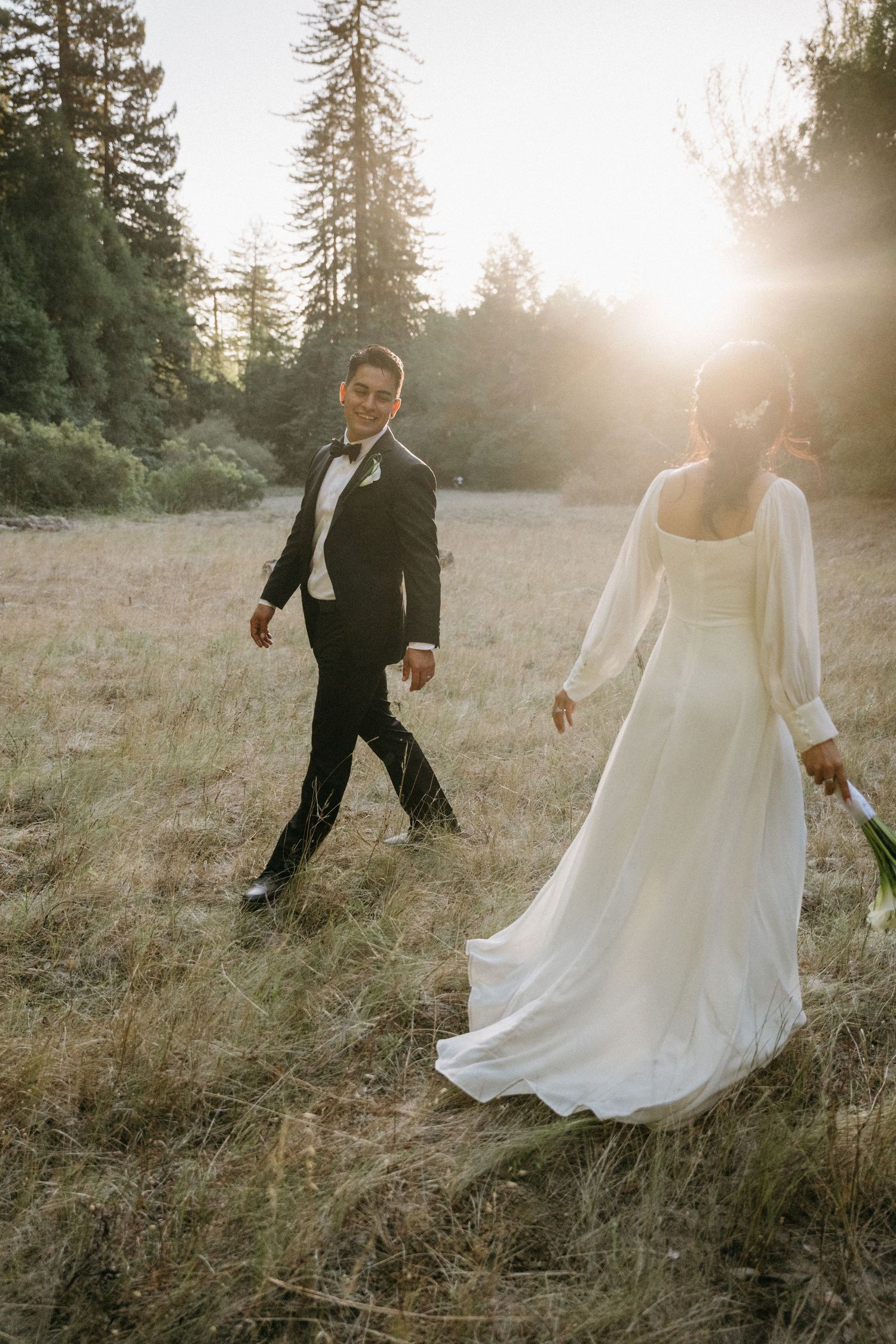 A bride in a flowing white gown and groom in a black tuxedo walk through a sunlit meadow surrounded by tall pine trees.