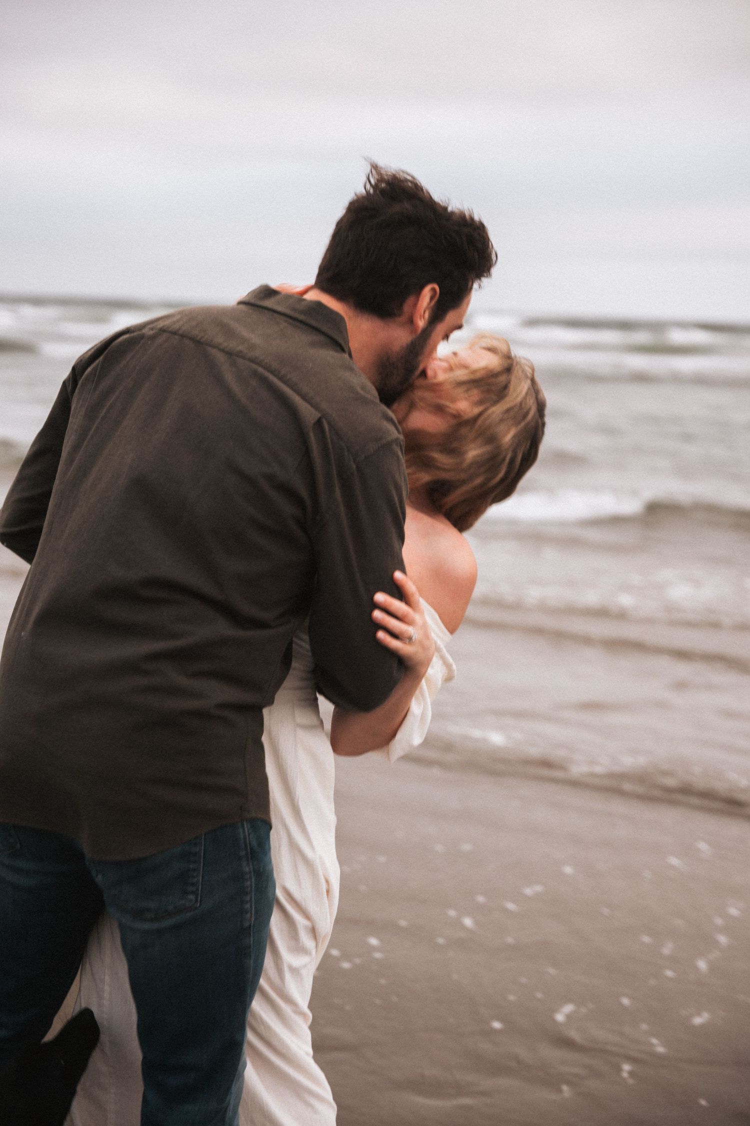 Couple sharing a romantic kiss on a windy beach, man in dark shirt embracing woman in white dress by the ocean.