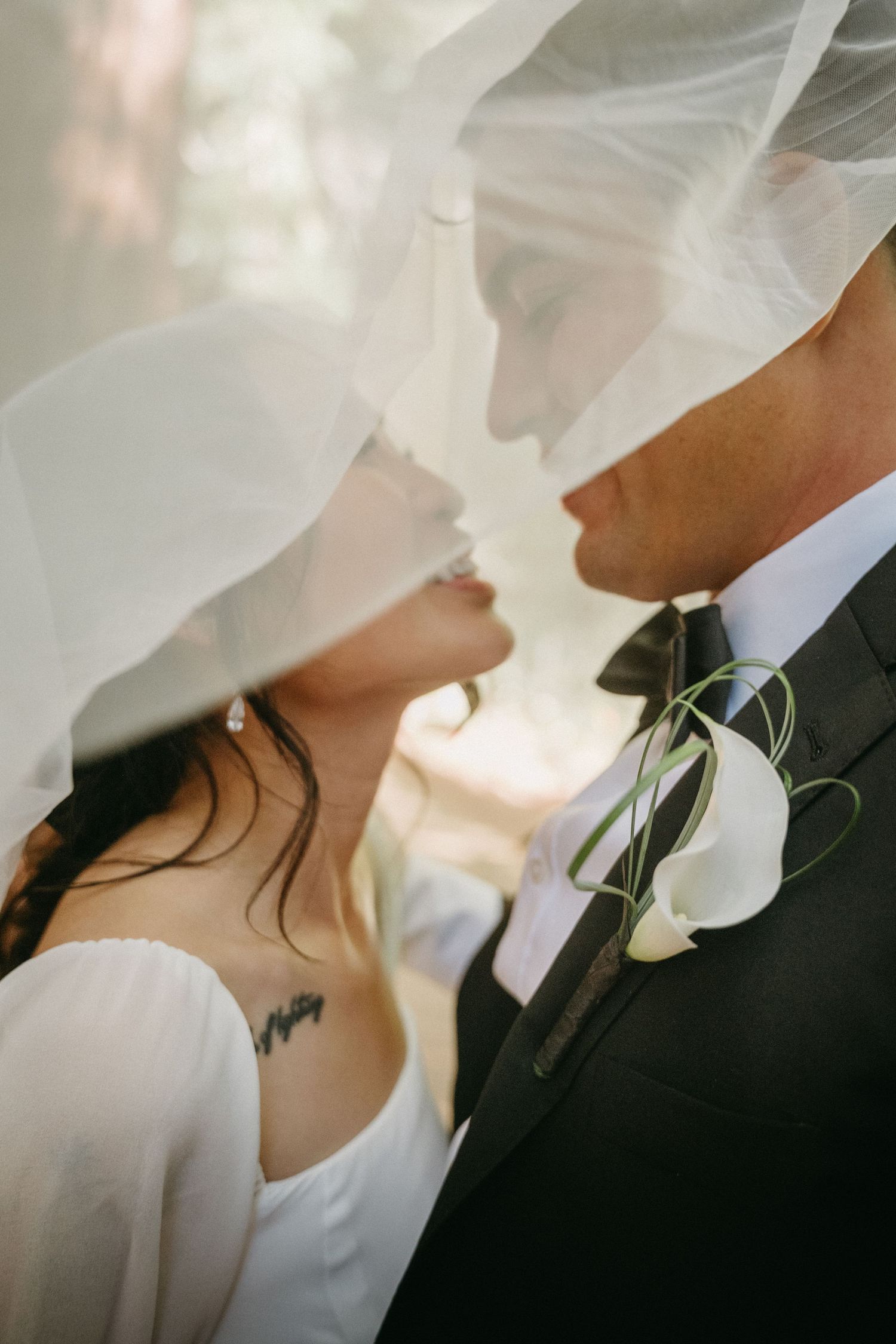 Bride and groom sharing an intimate moment under a wedding veil, groom wearing a black suit with white calla lily boutonniere.