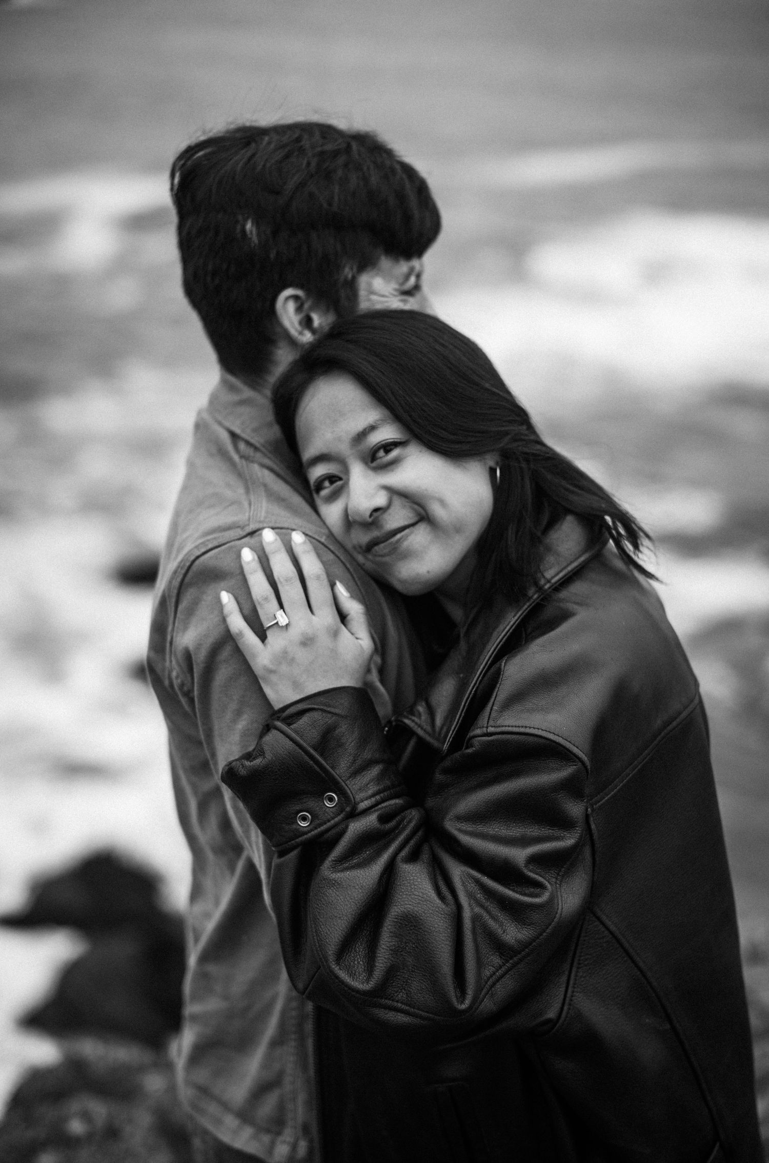 Black and white photo of a couple embracing near the ocean, woman in leather jacket smiling with engagement ring visible.