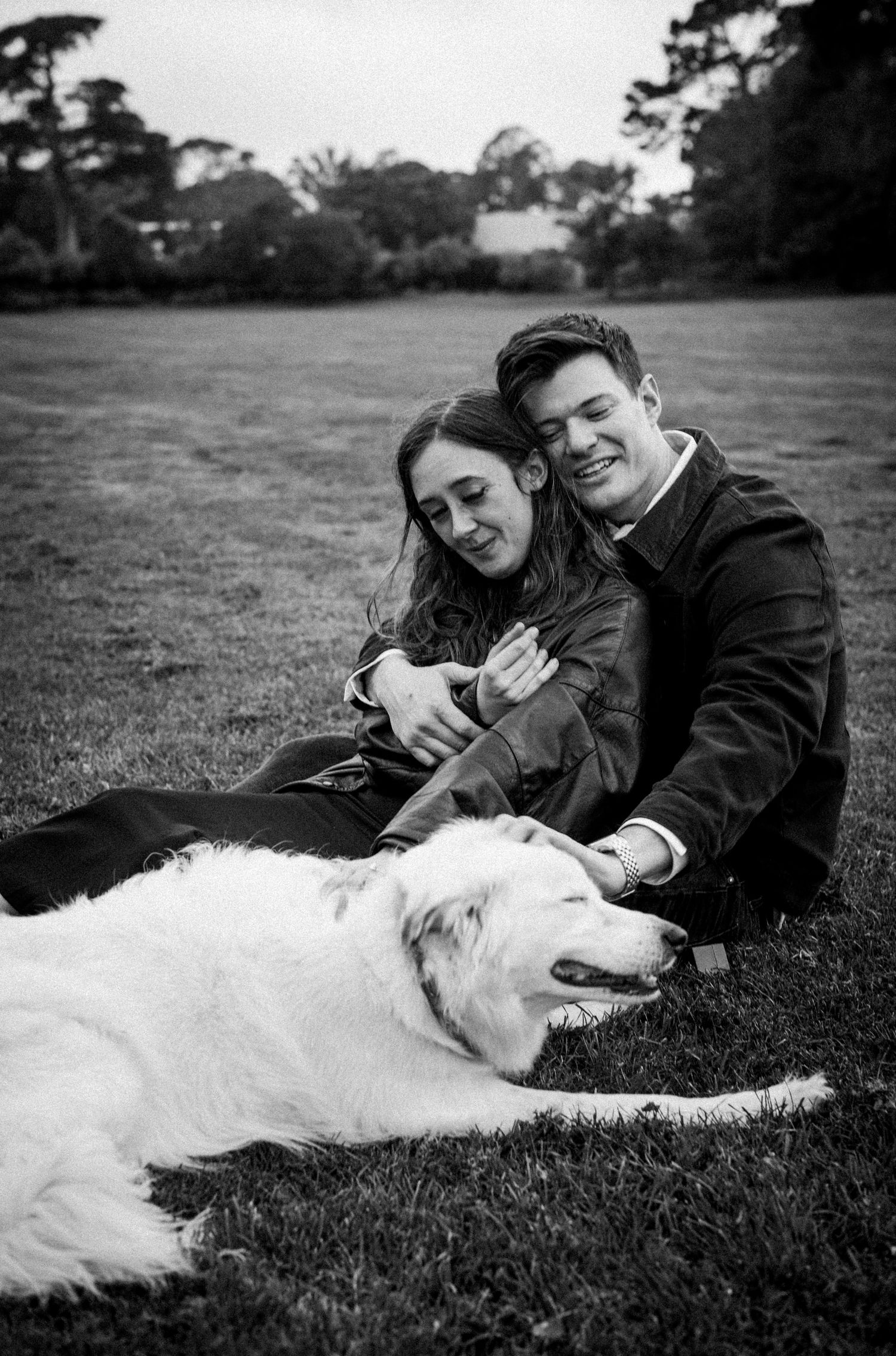 Black and white photo of smiling couple sitting on grass with their white fluffy dog.