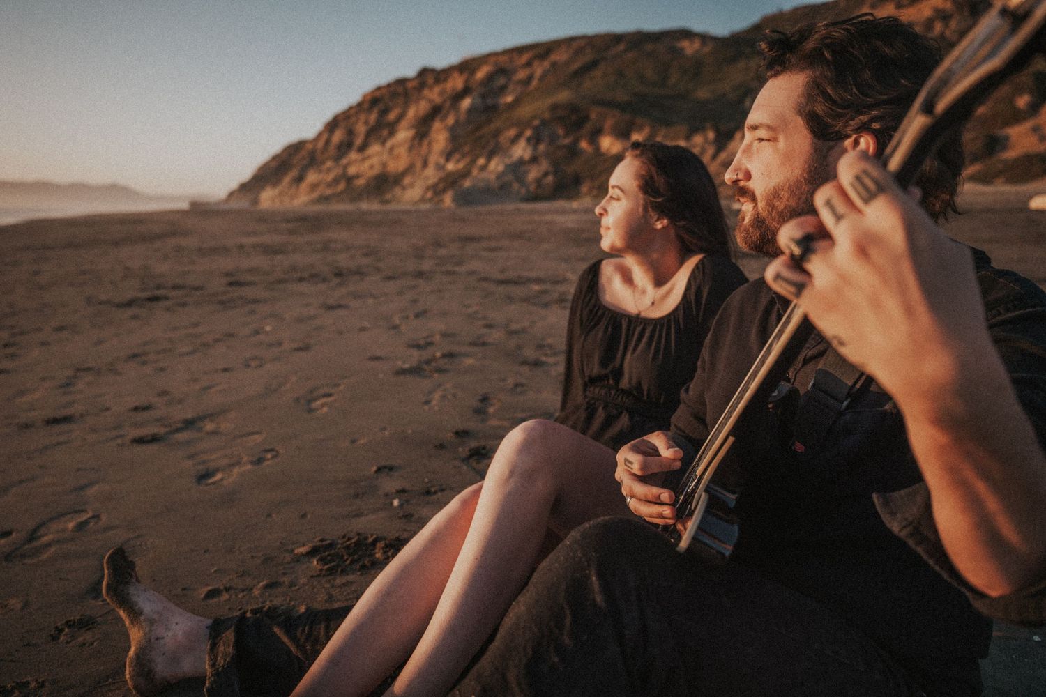 Couple sitting on sandy beach at golden hour, man playing guitar while woman gazes at ocean view.