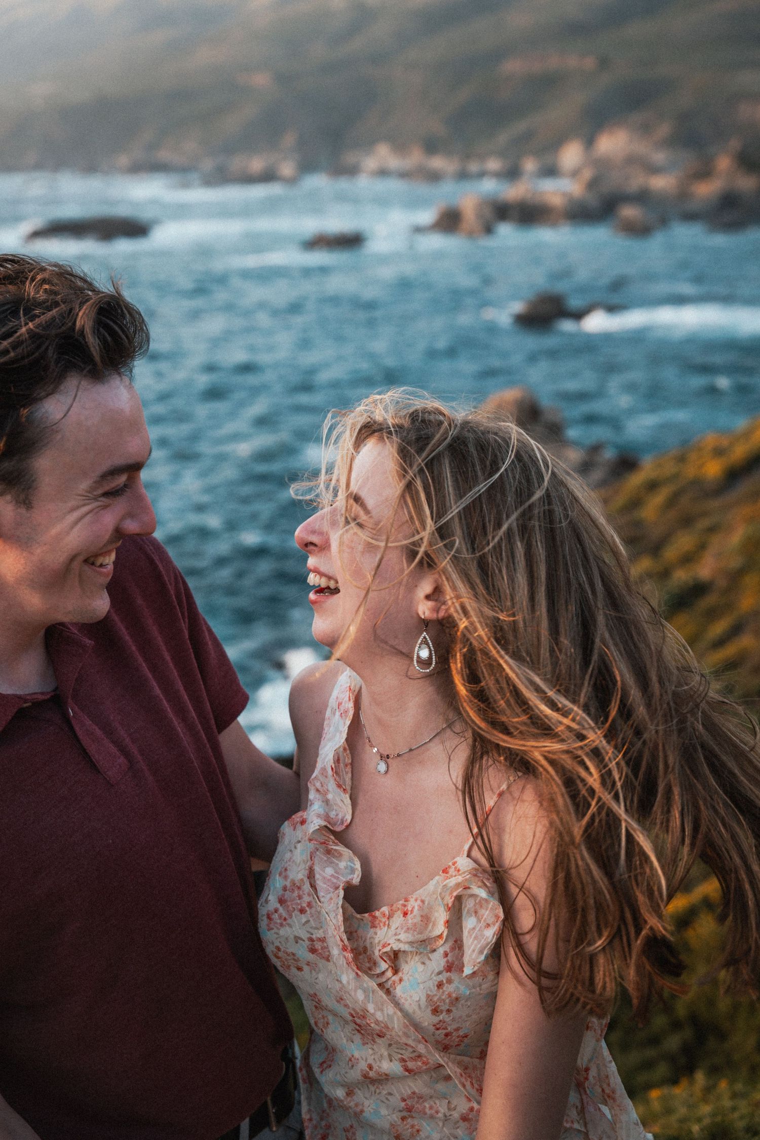 Laughing couple in floral dress and red shirt with rocky ocean coastline at sunset behind them.