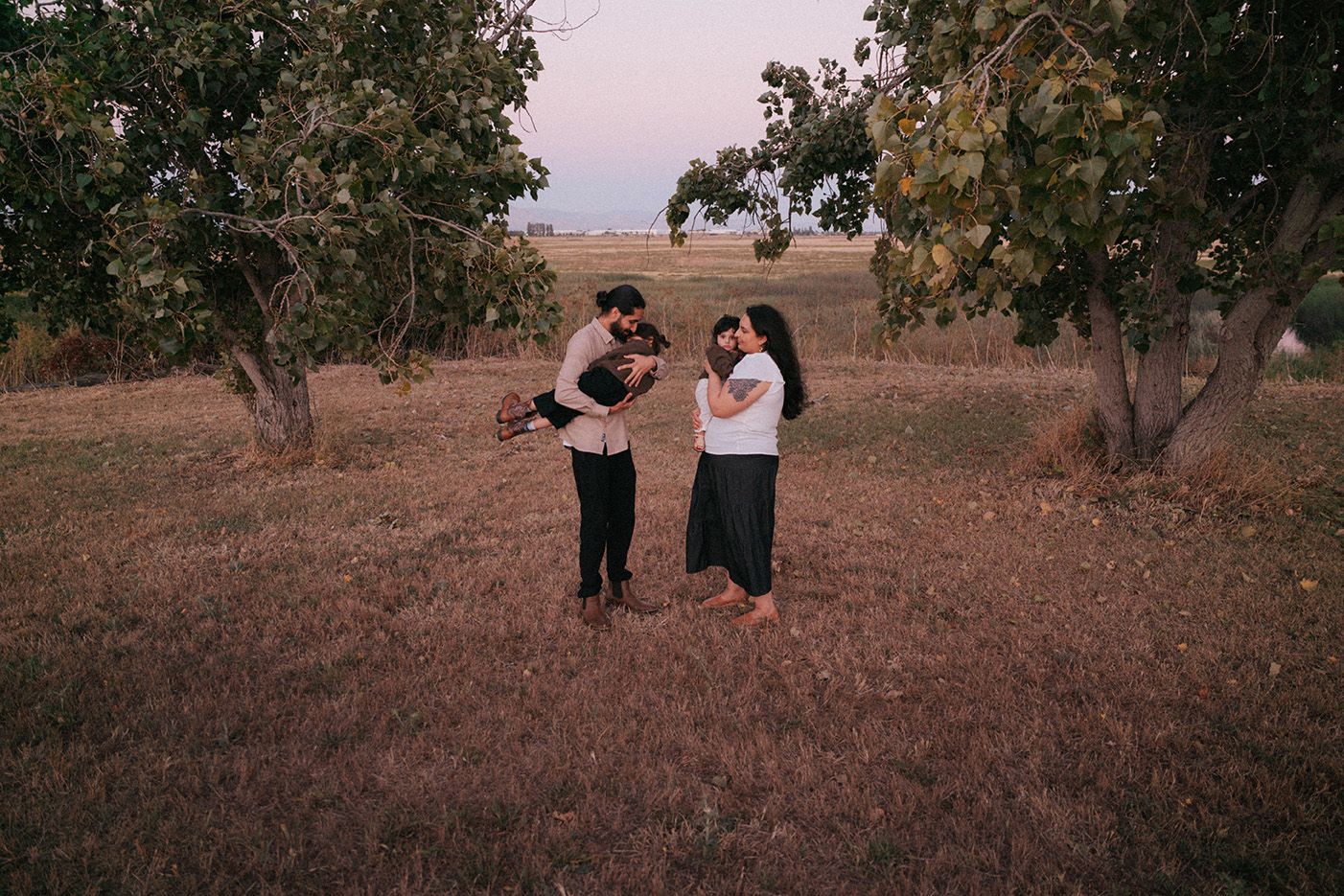 A family of four stands among trees in an open field at dusk, sharing tender moments in a serene outdoor setting.