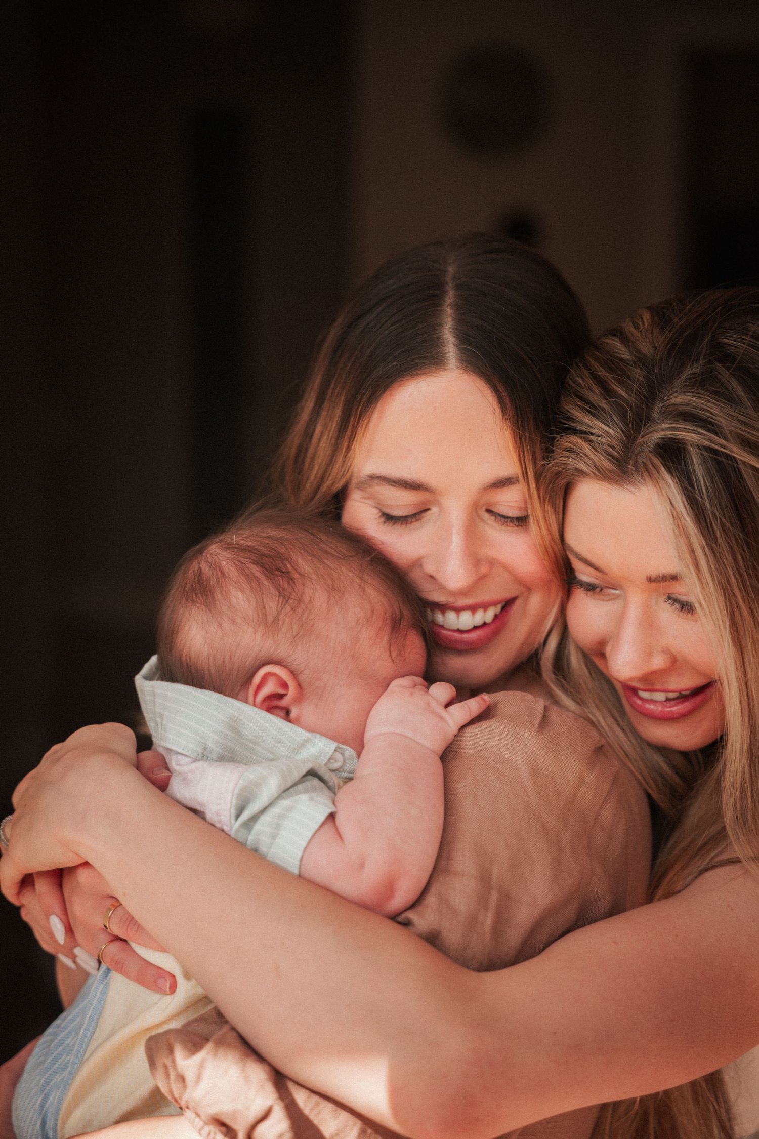 Two women smiling and embracing a newborn baby, sharing a tender family moment in warm natural light.