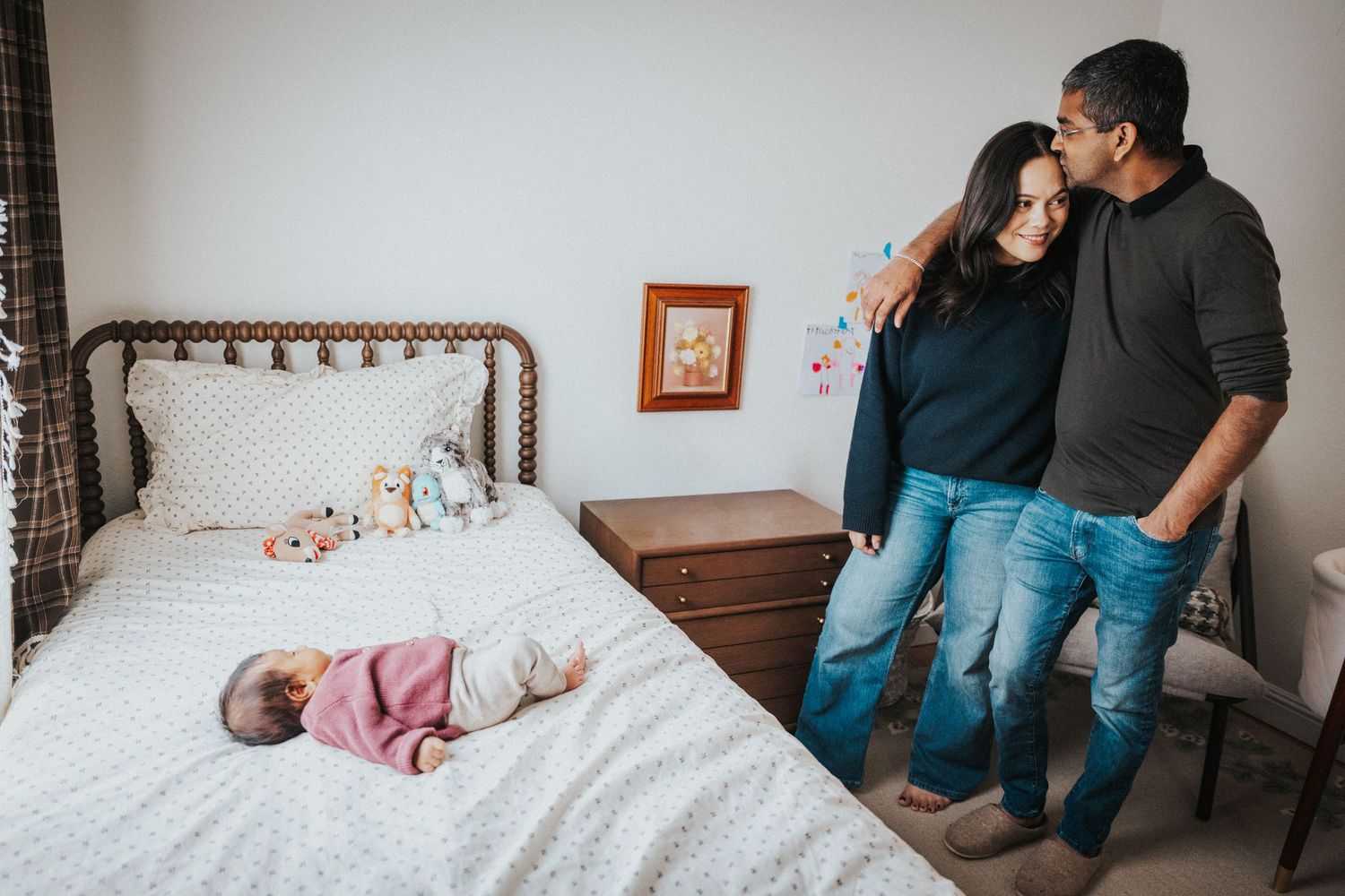 A couple stands embracing in a bedroom while their newborn baby lies on the bed beside them.