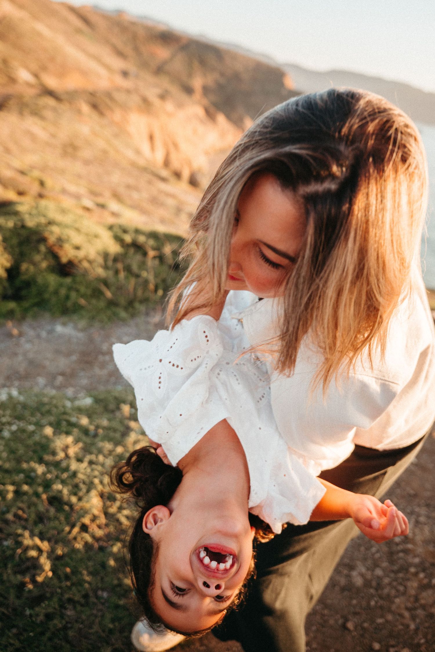 A joyful mother holds her laughing toddler upside down outdoors in golden sunlight with hills in the background.