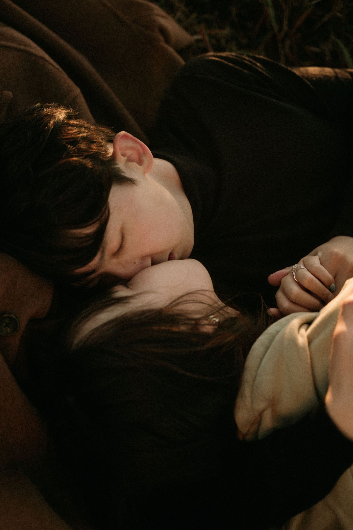 Romantic couple lying close together outdoors in warm golden light as a dark-haired man gently kisses a woman's forehead.
