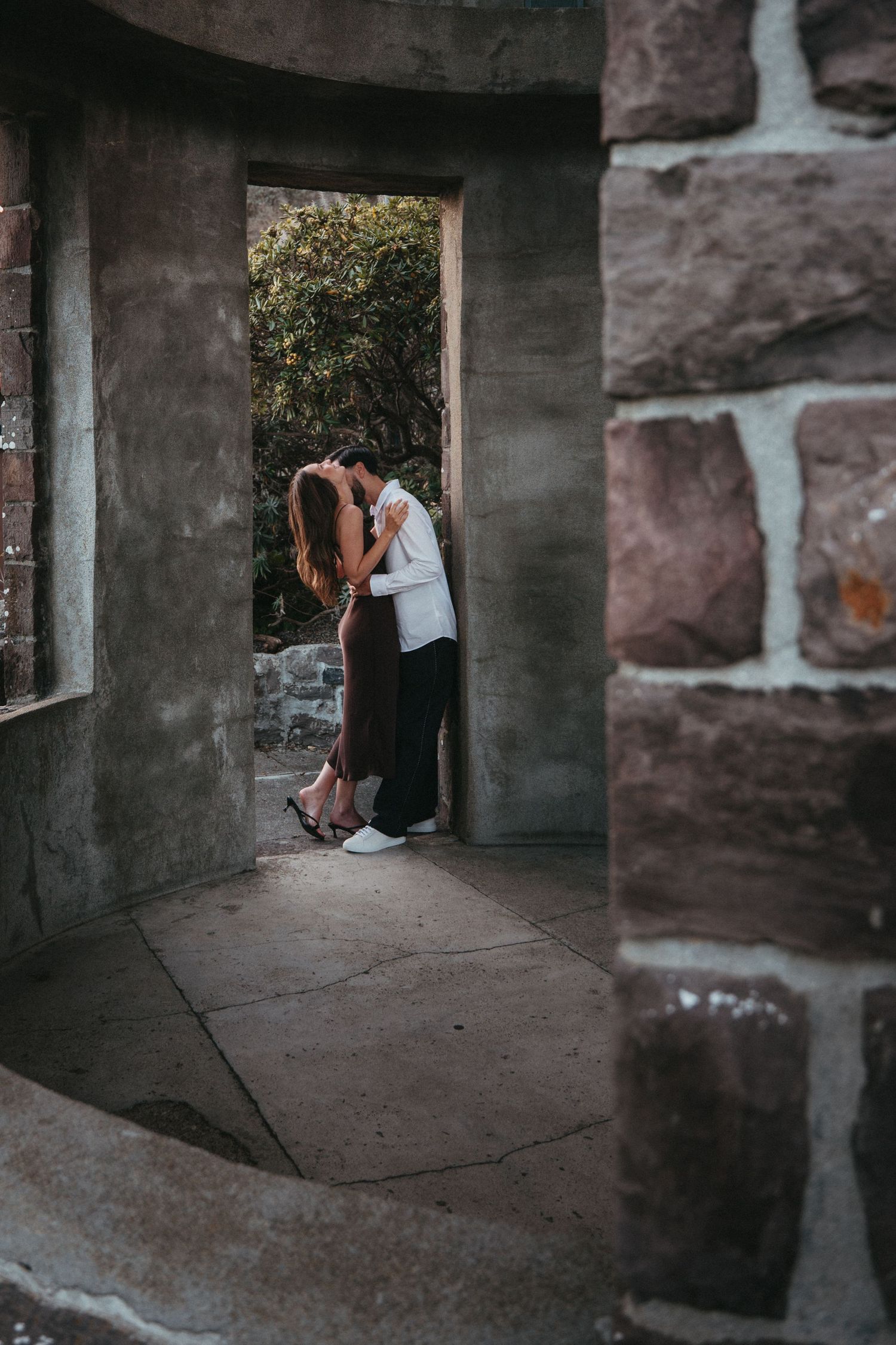 Couple embracing and kissing framed by a stone archway doorway in a rustic setting.