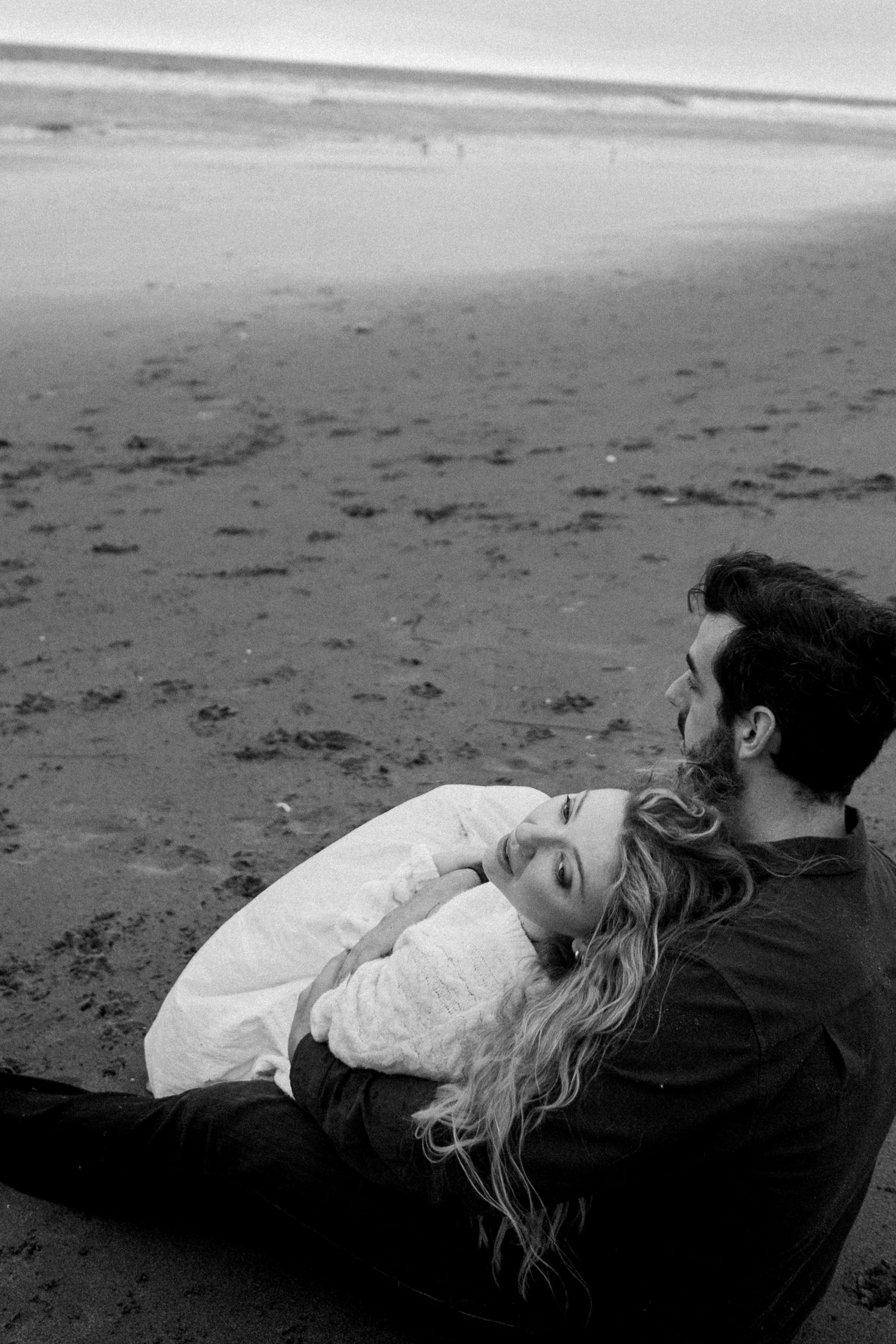 Black and white photo of a couple sitting together on a beach, woman resting head on man's chest.