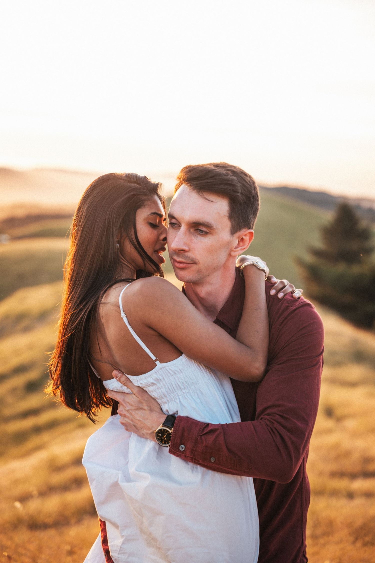 Couple embracing at golden sunset on a hillside, woman in white dress hugging man in burgundy shirt.
