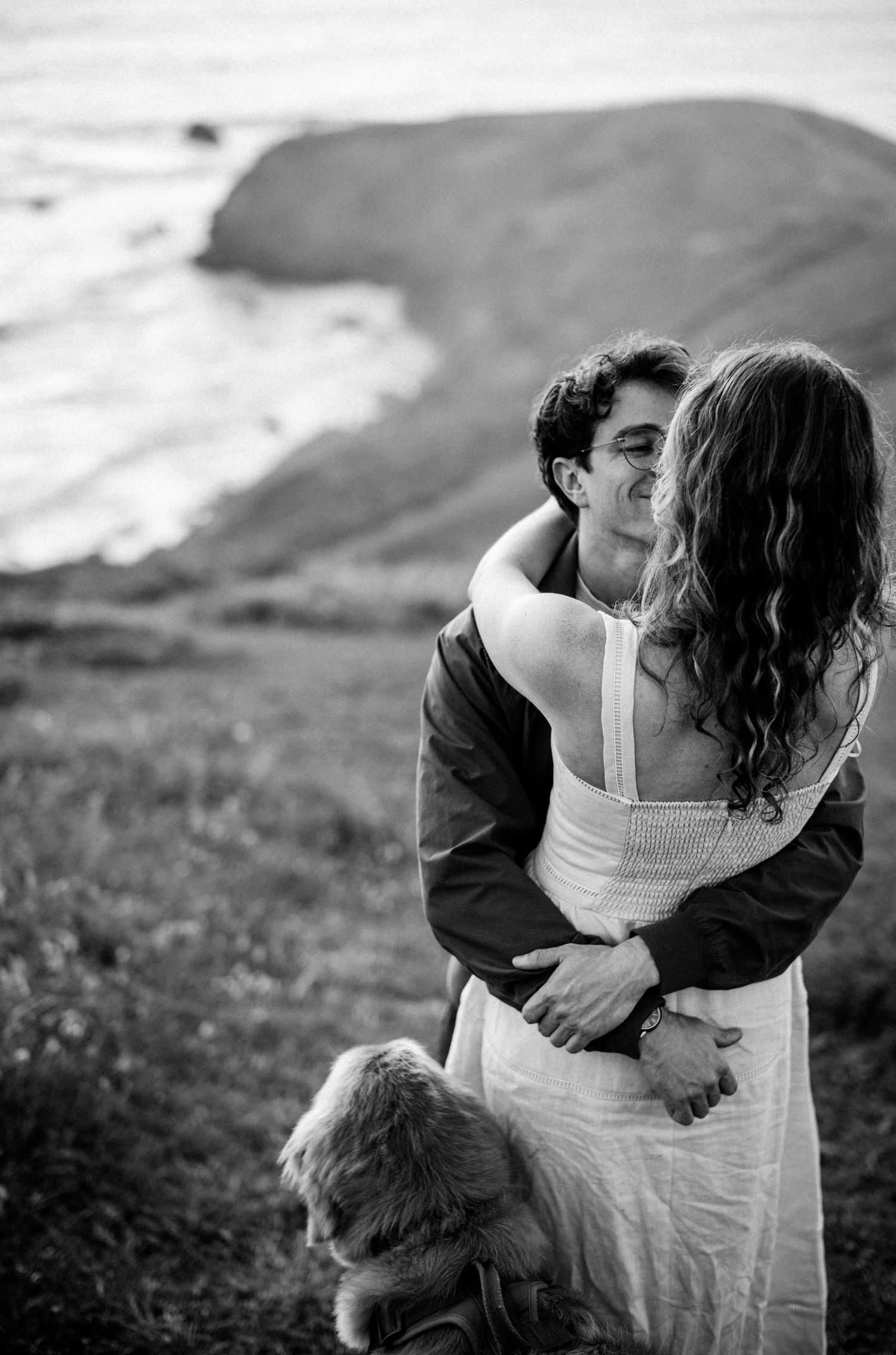 Man holds woman from behind as dog sits nearby on grassy coastal cliff in black and white photo.