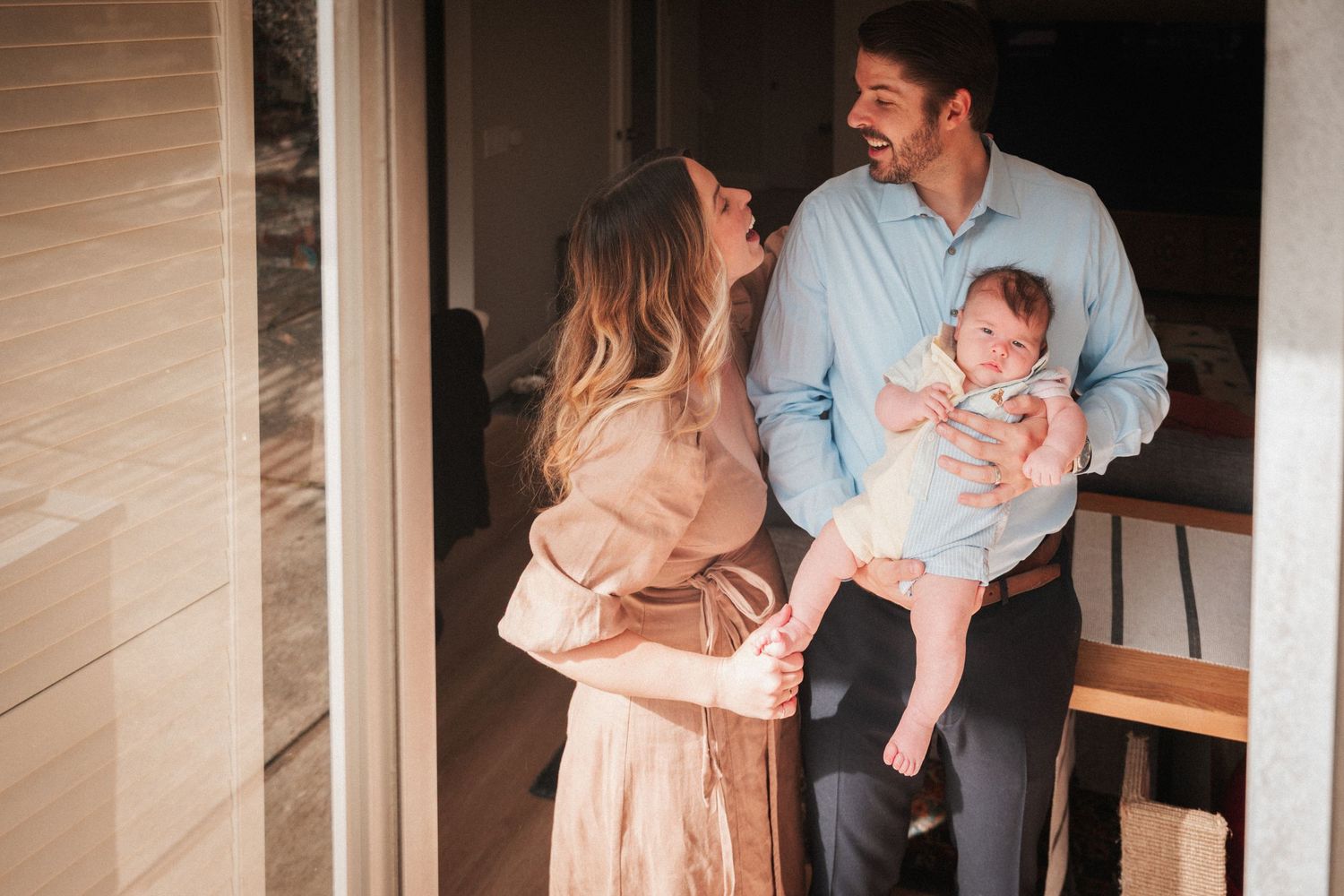 Happy young couple smiling at their newborn baby near a modern home entrance in warm natural light.
