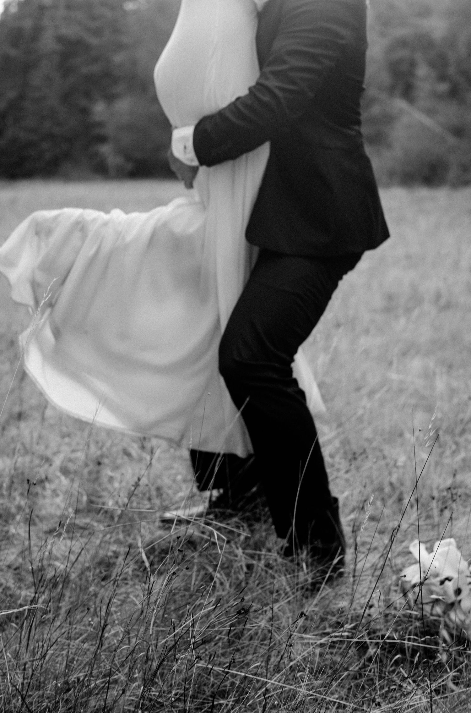 Black and white photo of bride and groom walking through a grassy field on their wedding day.