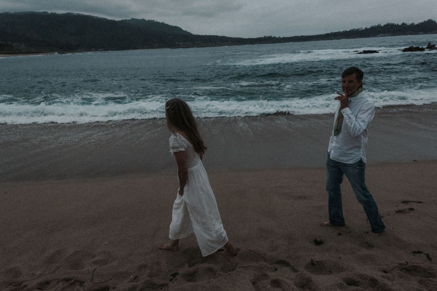 A couple in white clothing walks along a sandy beach with ocean waves and misty green hills in the background.Photography by The Y.K. Studio