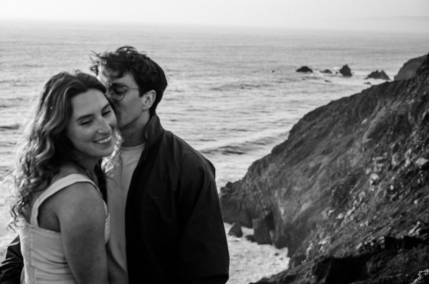 Black and white photo of a couple embracing on a rocky coastal cliff with ocean waves in the background. Photography by The Y.K. Studio.
