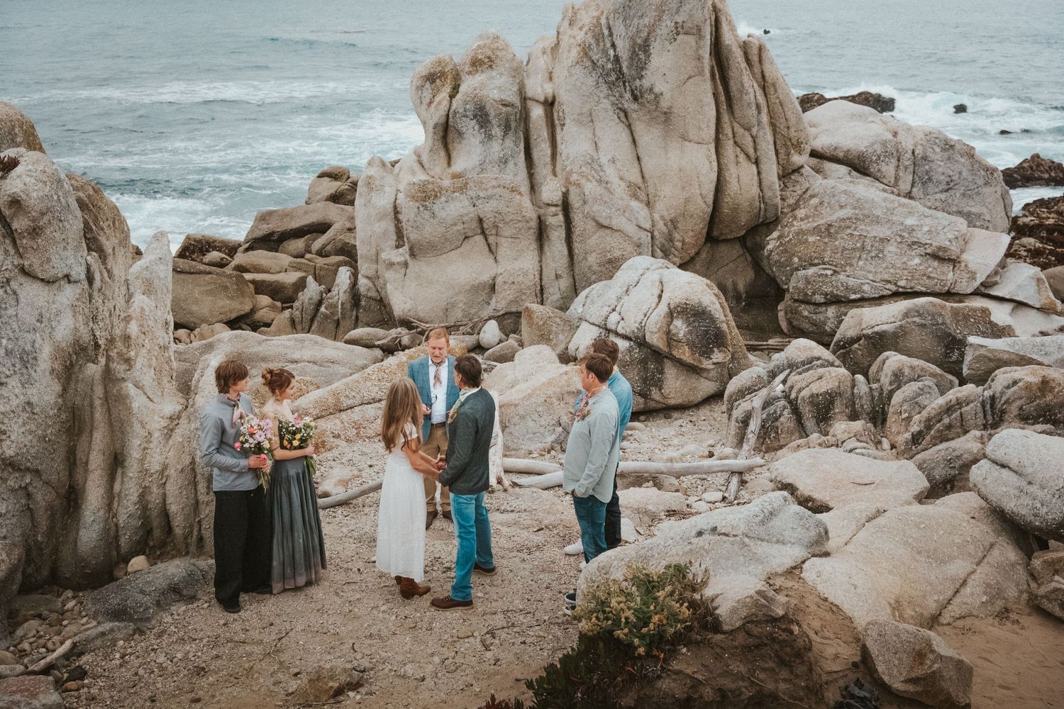 Small intimate beach wedding ceremony among dramatic coastal rocks with ocean backdrop.