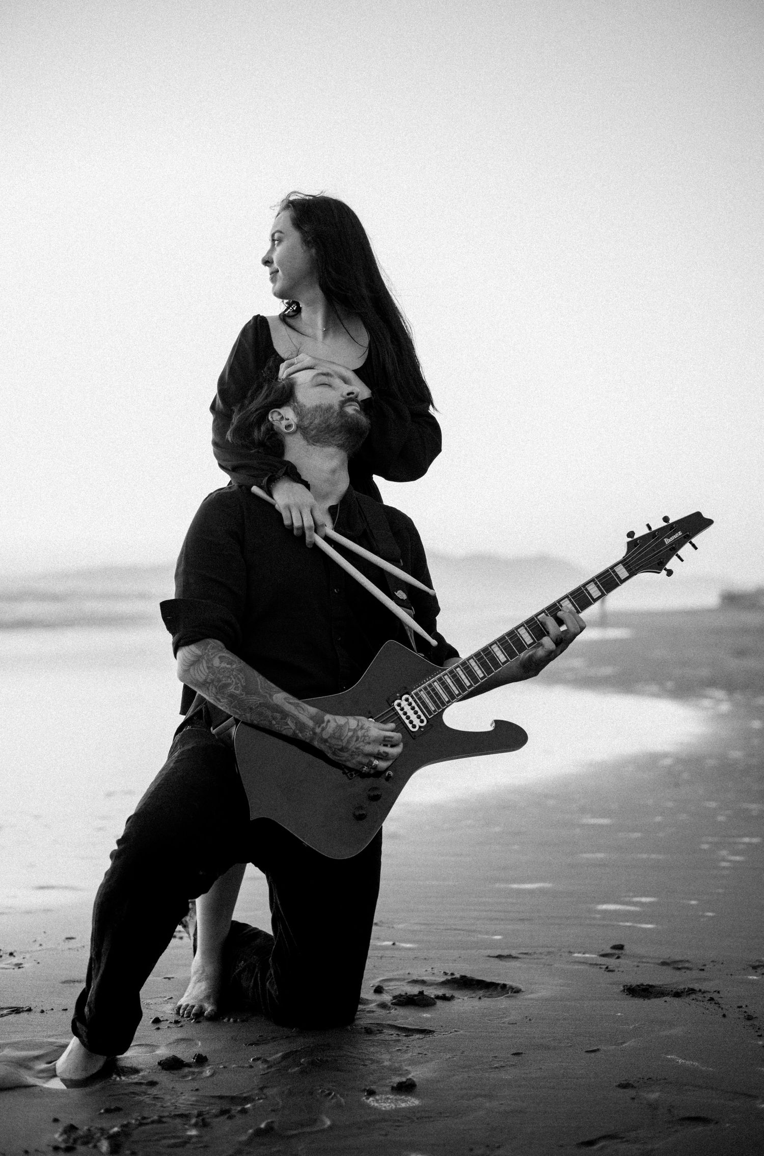 Black and white beach photo of a man kneeling with electric guitar while woman stands behind him.