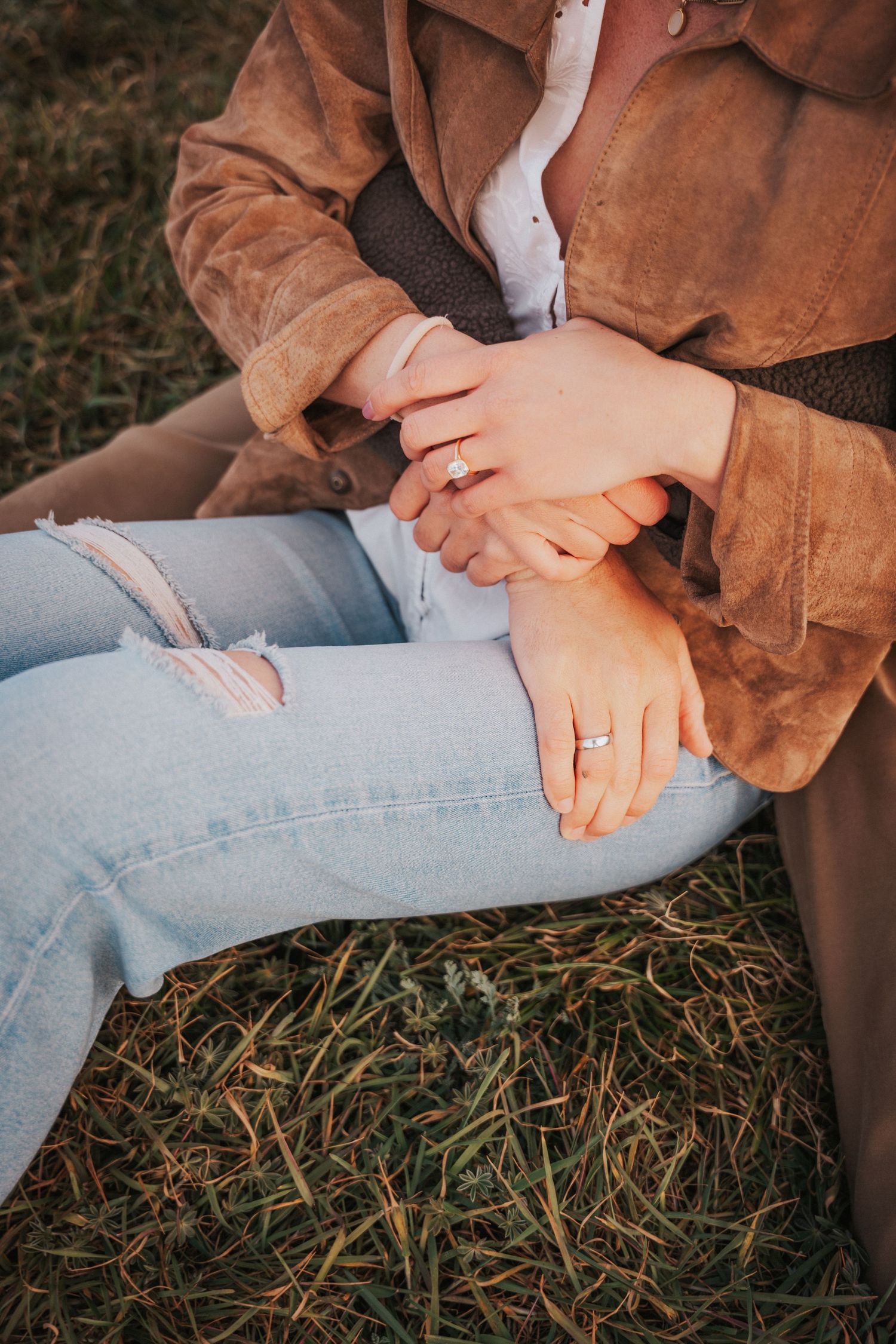 Couple holding hands outdoors, wearing rings, sitting on grass in jeans and brown suede jacket.Photography by The Y.K. Studio.