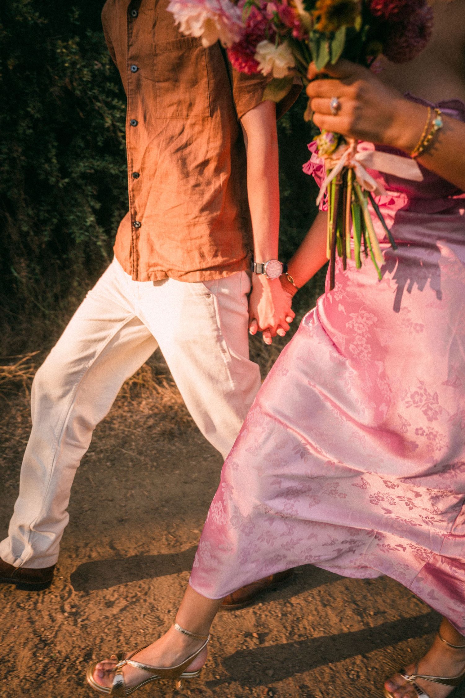 Couple walking hand in hand, woman in pink floral dress holding bouquet, man in brown vest and white pants.