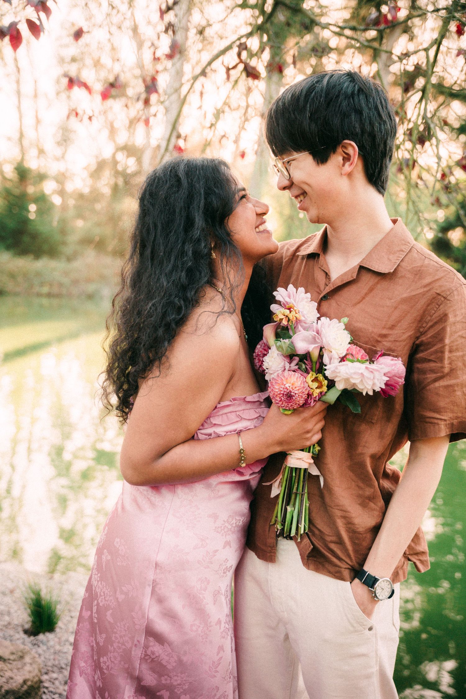 A romantic couple shares a tender moment outdoors, the woman in a pink dress holding a bouquet of pink flowers near a serene pond.