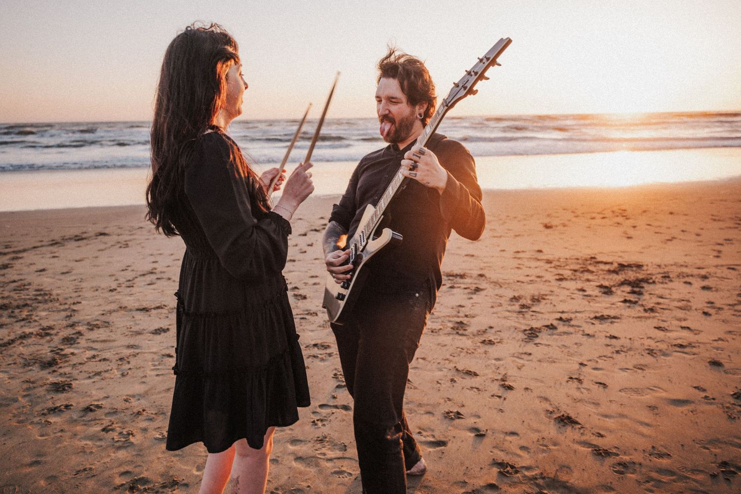 Man and woman laughing on sandy beach at sunset, she holds drumsticks, he plays electric guitar.