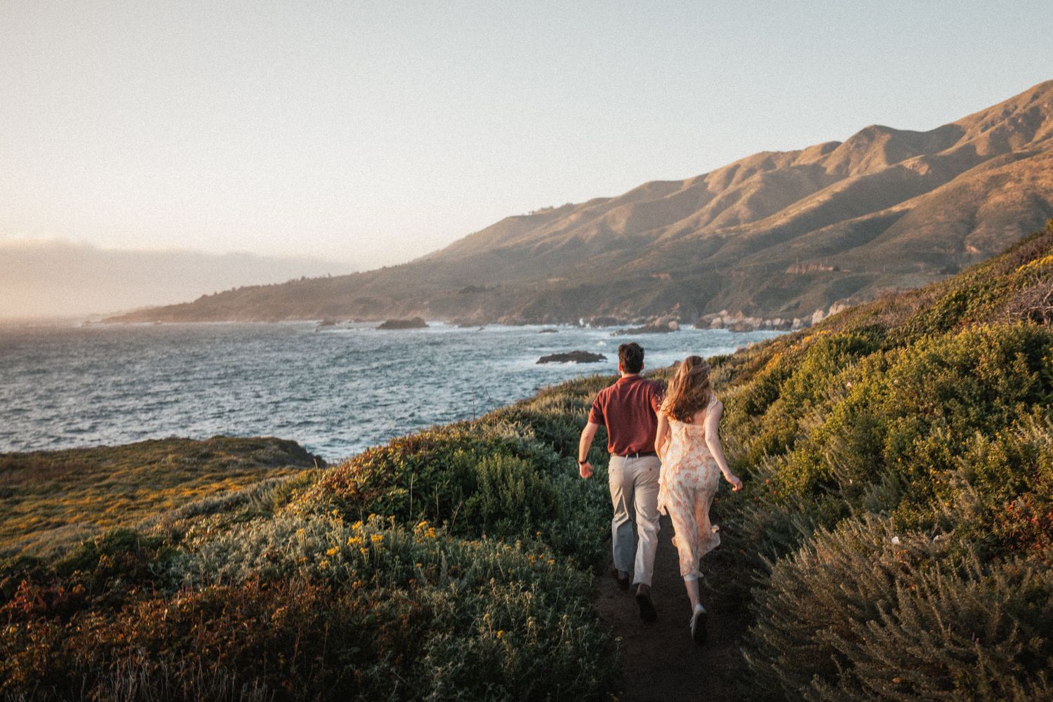 Couple walking along a scenic coastal trail with ocean views and golden hills at sunset.
