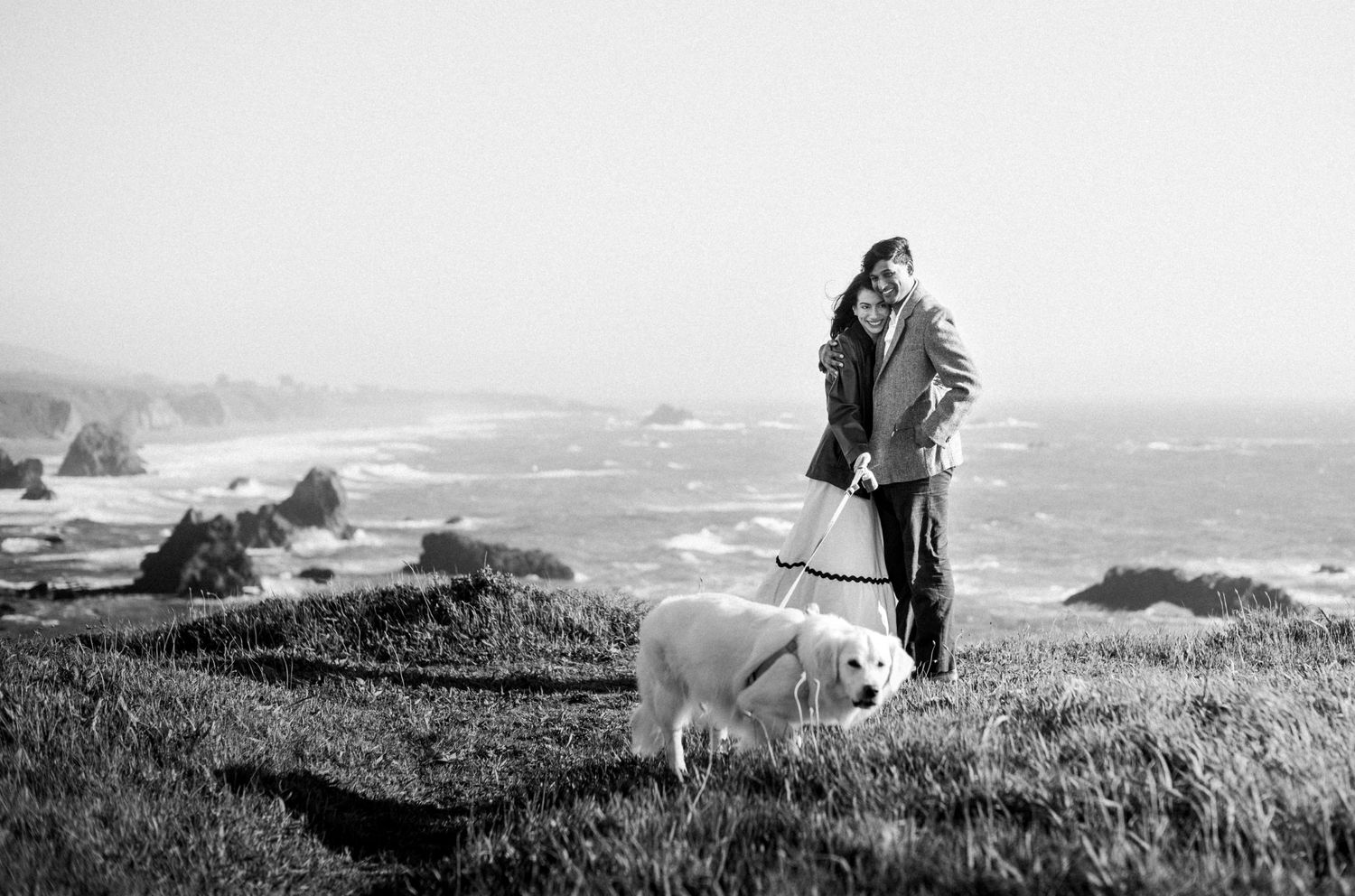 Couple walking white dog on grassy coastal bluff, black and white photo, misty ocean view.