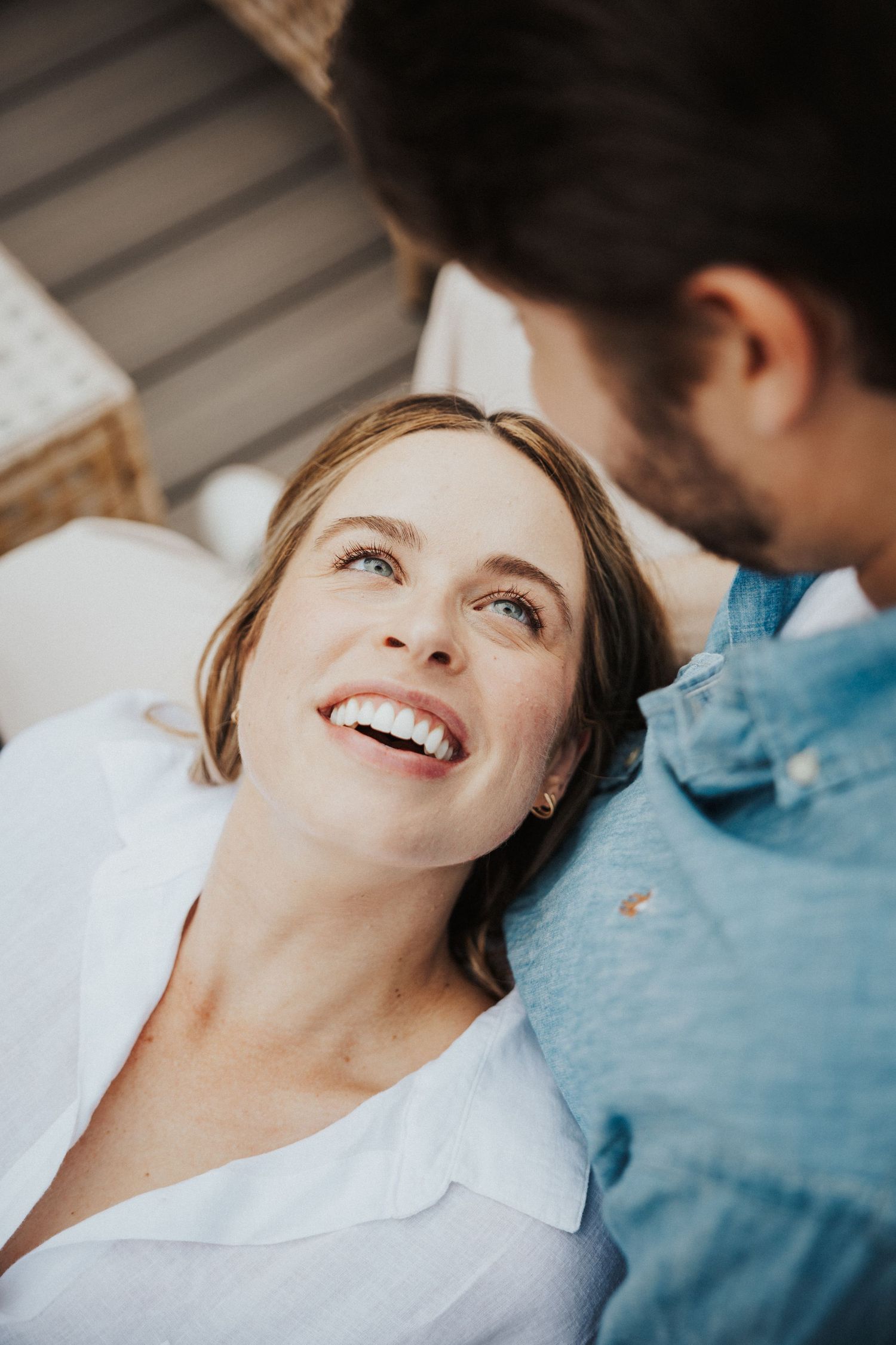 A smiling woman in a white shirt looks up at a man in a blue denim shirt in a warm, intimate moment.