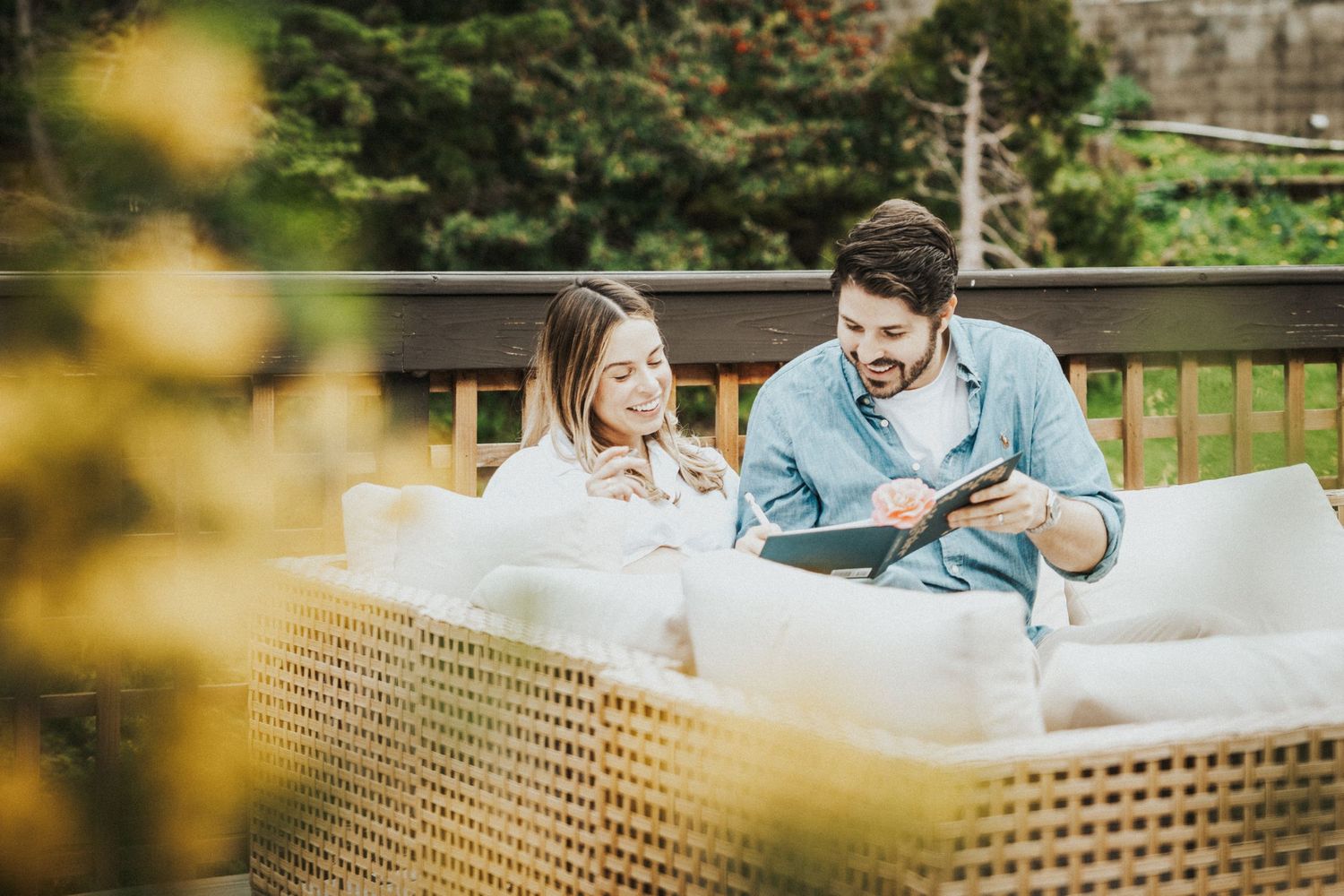 Couple smiling and reading a book together on an outdoor patio sofa surrounded by lush greenery.