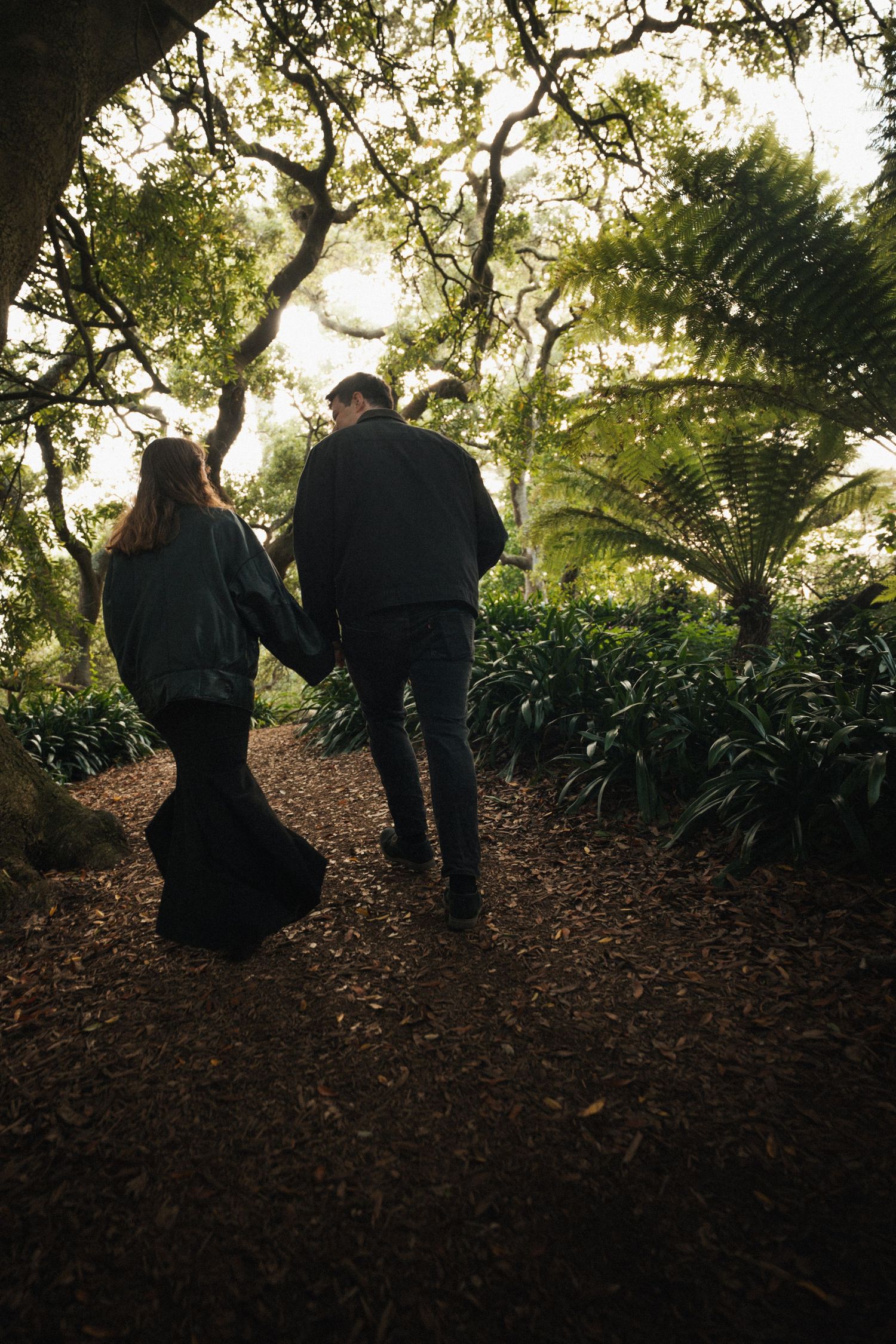 Couple holding hands walking through a lush green forest garden path covered in brown mulch.