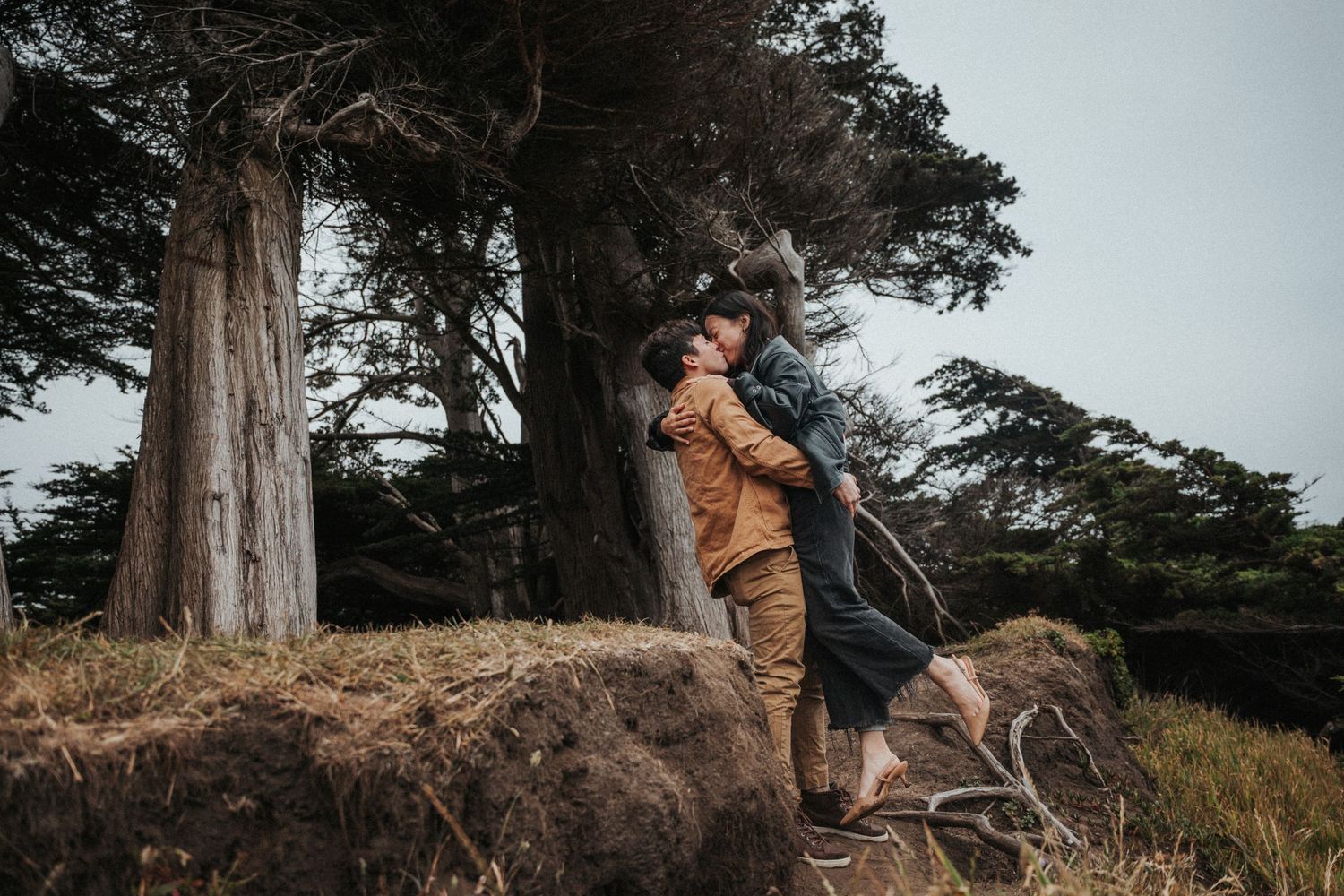 Couple embracing passionately near tall trees in a misty outdoor landscape.
